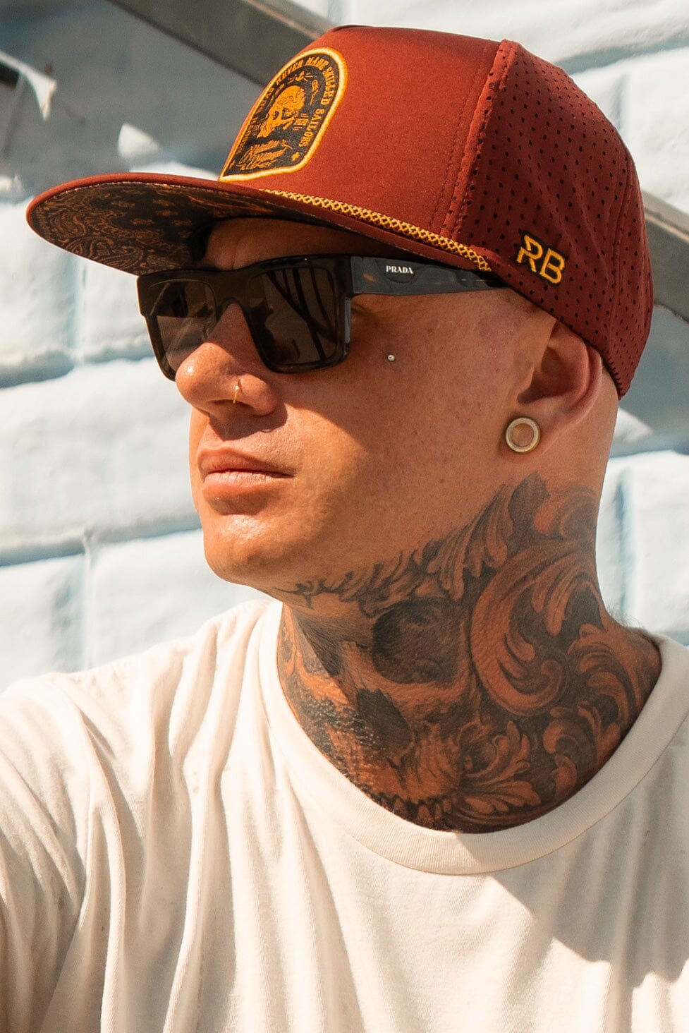 A person is outdoors against a light-colored brick wall, wearing the Relentless Betrayal Skilled Sailor Snapback in burgundy, large black sunglasses, and a white shirt. They have ear gauges, a nose piercing, and neck tattoos while looking to the side in their water-resistant cap.