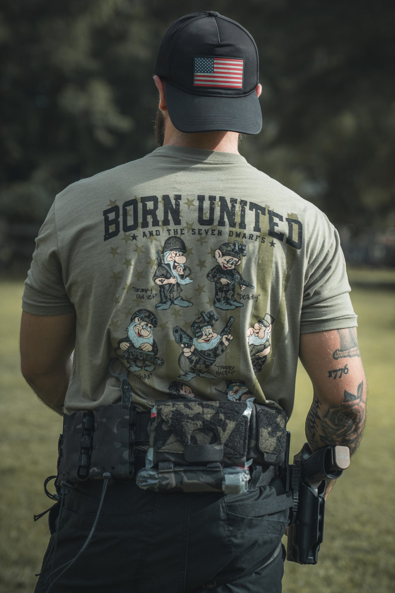 A man stands outdoors with his back to the camera, wearing a Born United Private Label Seven Soldiers shirt featuring cartoon dwarfs, an American flag cap, tattoos, and a tactical belt with gear and a pistol.
