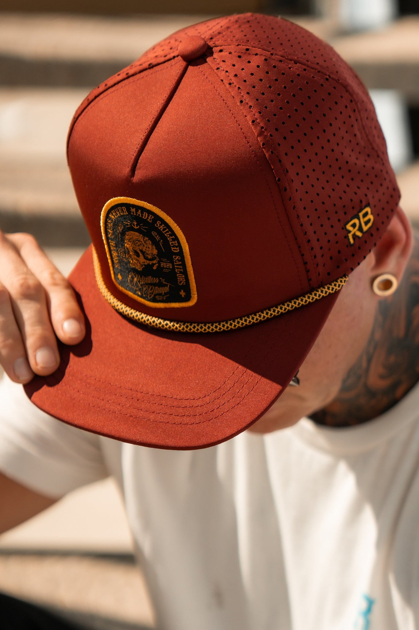 Wearing the Skilled Sailor Snapback - Burgundy from Relentless Betrayal, someone covers their face with the perforated handmade cap featuring a circular patch and gold chain. They also sport a white shirt with a tattoo and wooden ear plug.