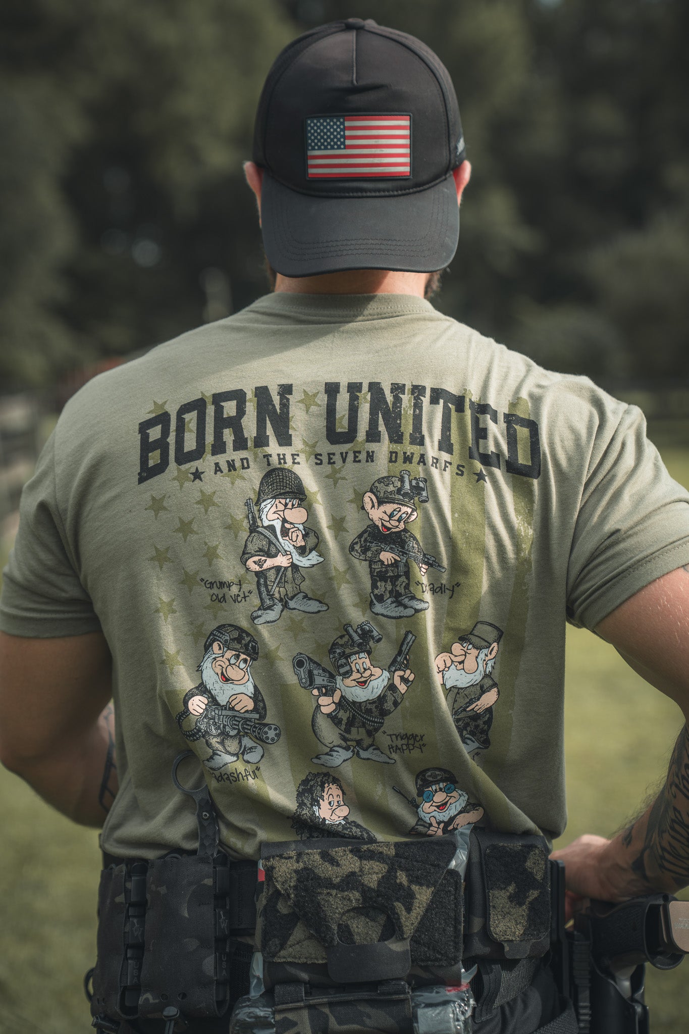 Wearing a Born United Private Label Seven Soldiers olive-green shirt with cartoon soldiers styled as dwarfs and a US flag cap, this person also sports tactical gear on their belt.