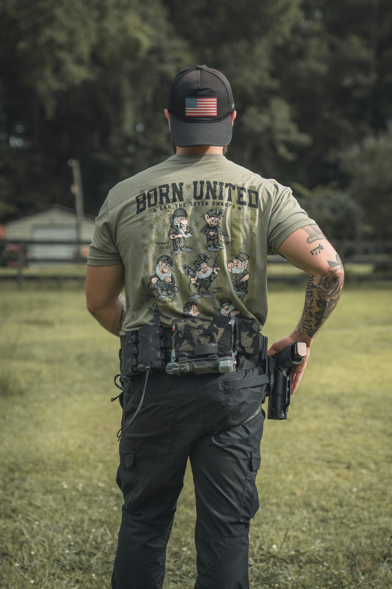 A man stands outdoors with his back to the camera, wearing a Born United Private Label Seven Soldiers green shirt with animated soldiers, an American flag cap, tactical belt, and visible arm tattoos.