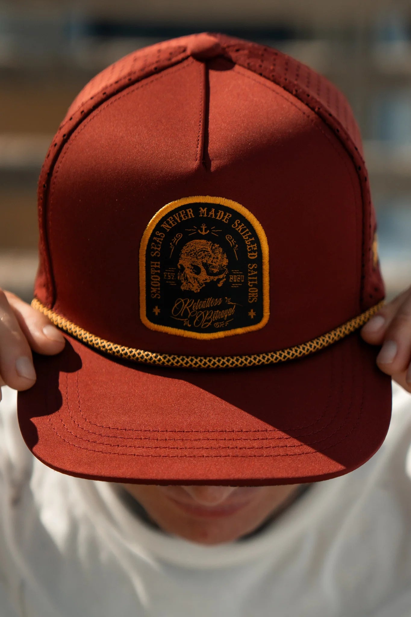 Wearing the Skilled Sailor Snapback - Burgundy from Relentless Betrayal, complete with a yellow emblem and text reading Isolation: A Haggard Life and The Greatest Sales Never Made, this handmade cap offers unique charm as the person grips its brim with both hands.