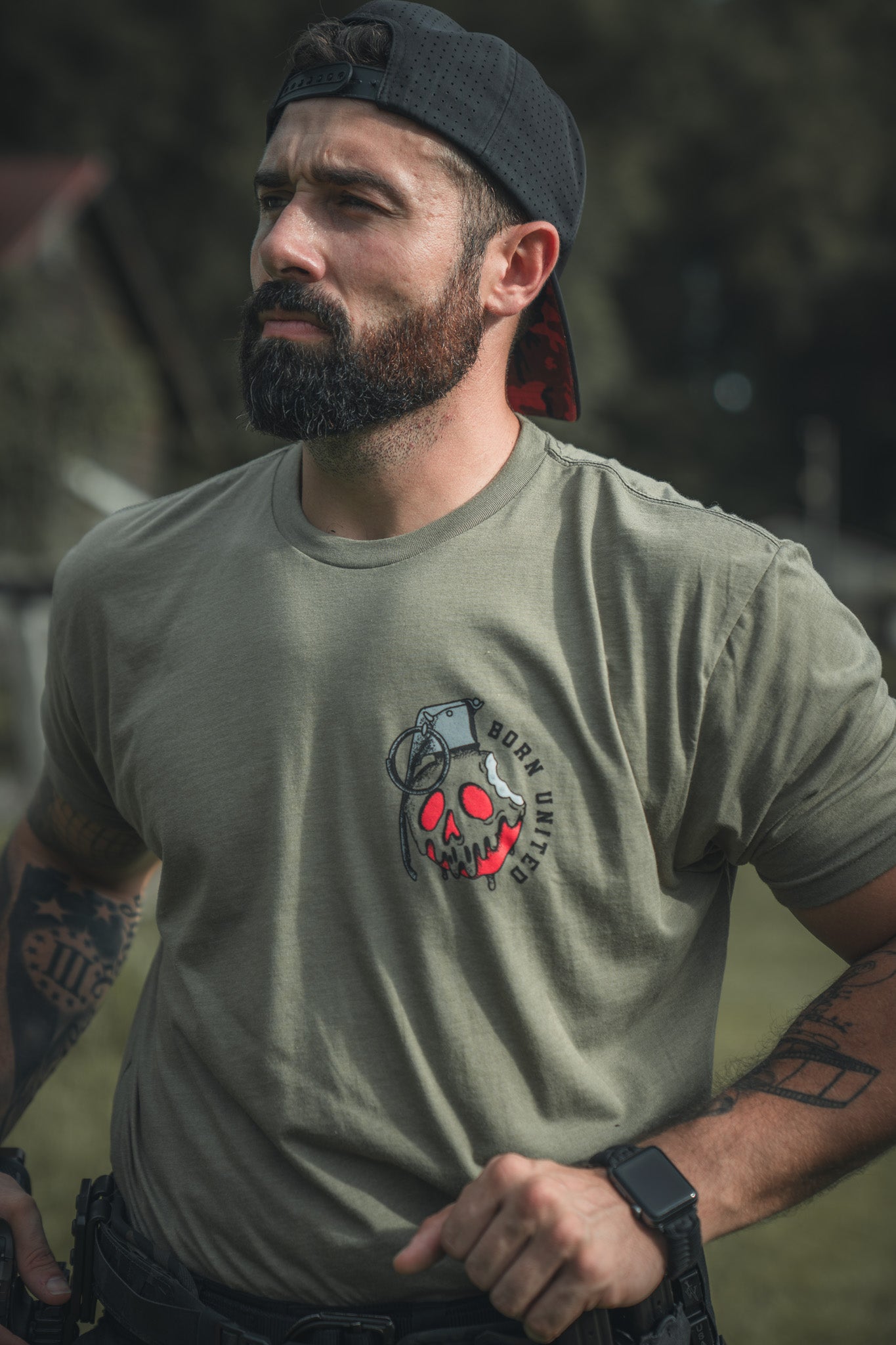 A bearded man in a backward black cap and olive green Seven Soldiers t-shirt by Born United Private Label stands outdoors, hands on hips, showing off his arm tattoos for a rugged, comic-inspired look.