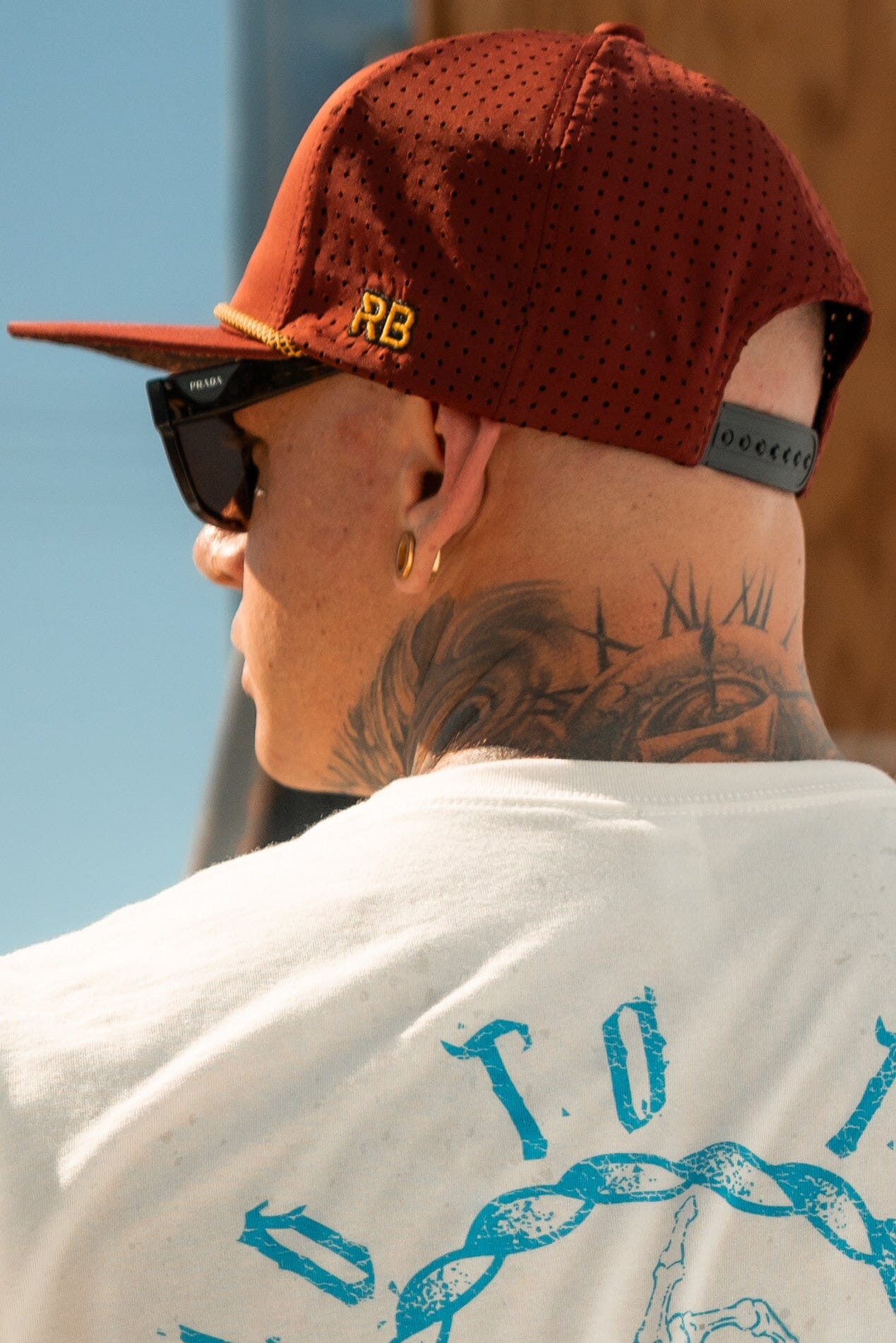 A person in a red, handmade hat and sunglasses faces away. Tattoos adorn their neck, and they wear a white T-shirt with blue graphics. The Skilled Sailor Snapback by Relentless Betrayal, detailed with RB, adds flair. A sunny background softly blurs behind this stylish look.