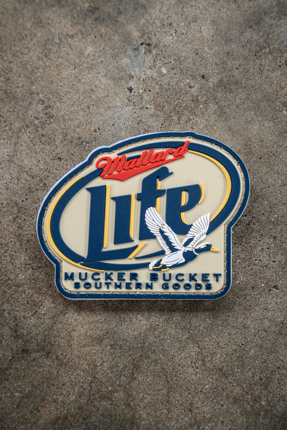 A Mallard Life PVC Patch by Mucker Bucket features a blue and red badge on concrete with a flying duck image and the text Mallard Life. Below, Mucker Bucket Southern Goods appears in blue letters.
