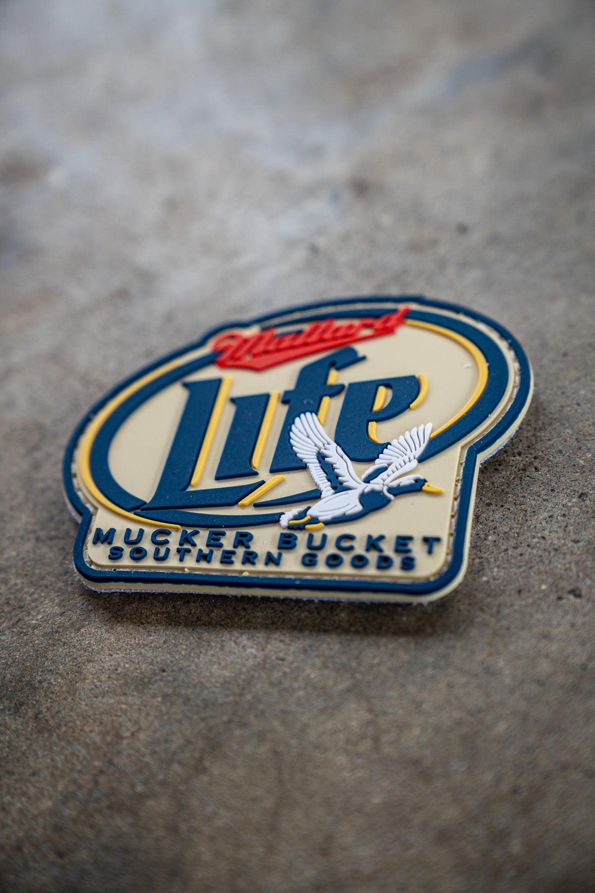 The Mallard Life PVC Patch by Mucker Bucket, featuring Mallard Life in large letters and two white birds near the bottom, sits on a textured surface; its design resembles the Miller Lite beer logo.