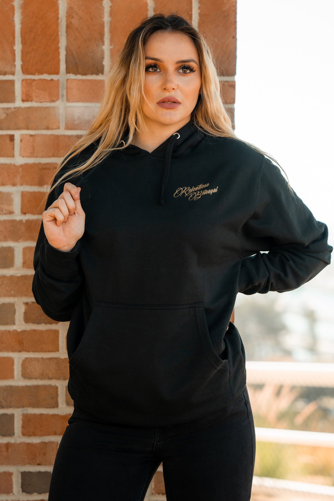 A blonde woman stands against a brick wall in jeans and a Skilled Sailor Gold Foil Hoodie by Relentless Betrayal. Her left hand is in her pocket, while her right holds the hoodie string, glancing slightly to the left.