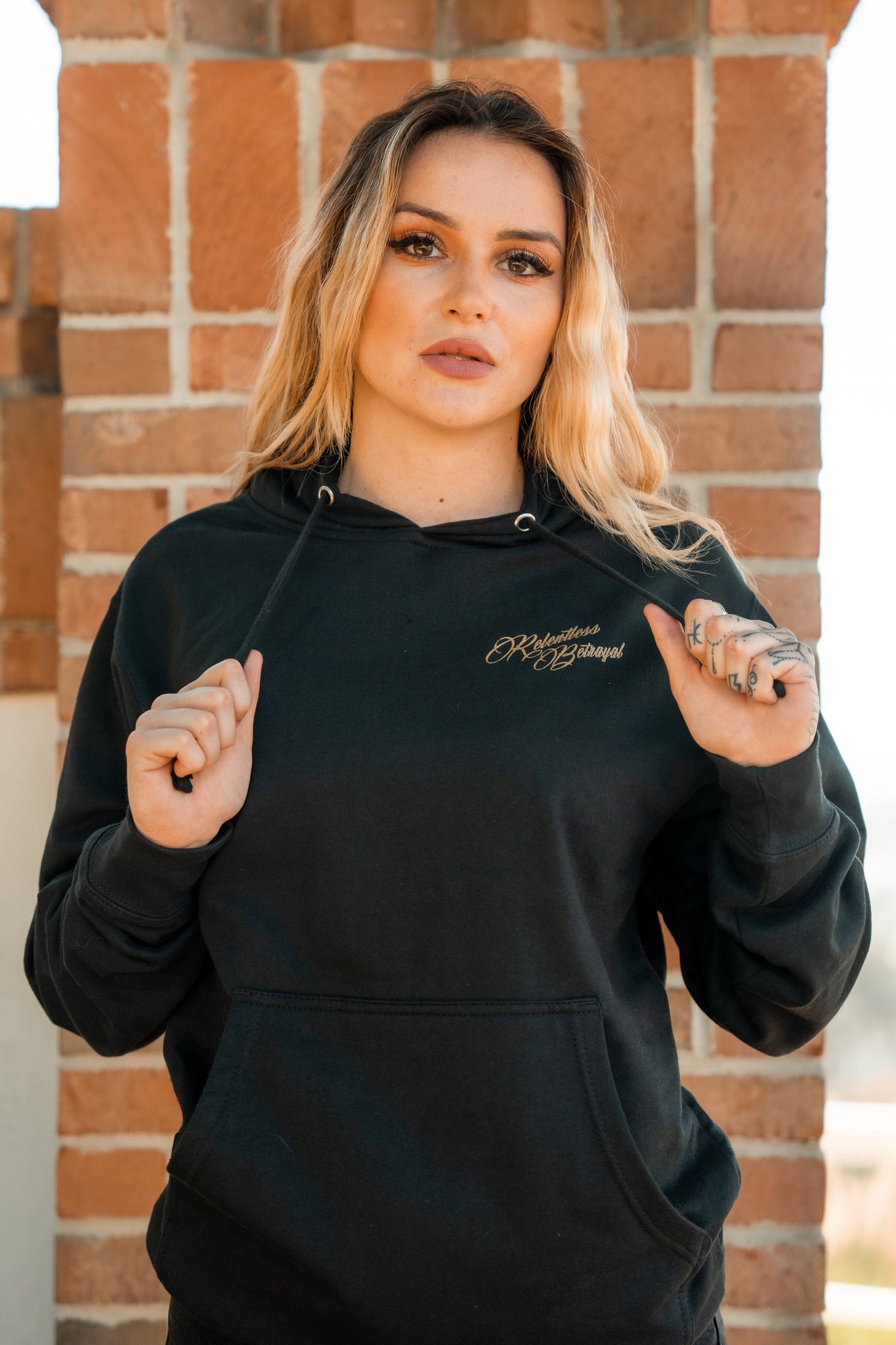 A person with long, wavy blonde hair wears the Skilled Sailor Gold Foil Hoodie by Relentless Betrayal. This unisex cotton/polyester hoodie combines comfort and style as they hold the strings, standing before a brick wall.