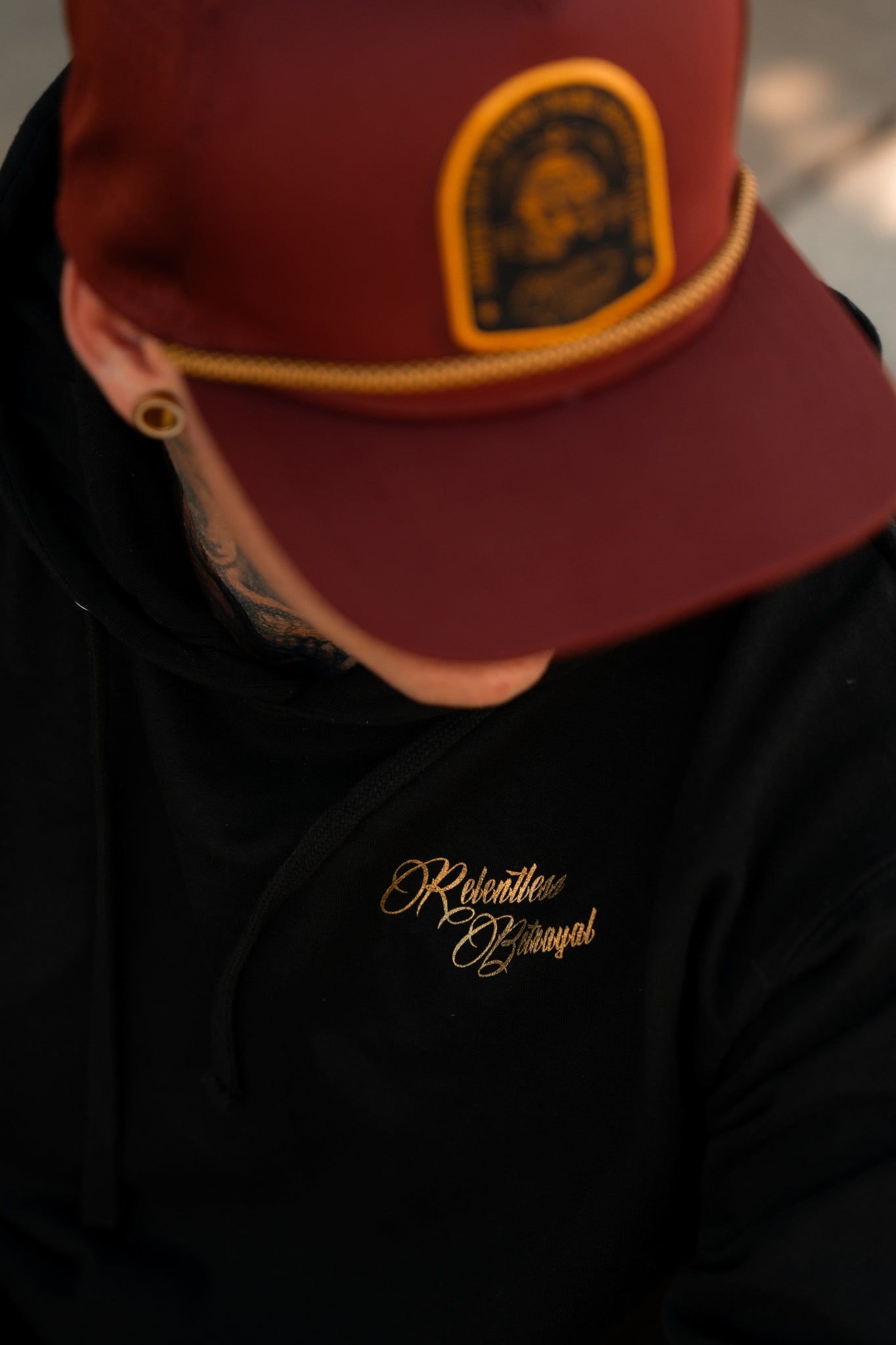 A person in a maroon cap with an embroidered patch and a Relentless Betrayal Skilled Sailor Gold Foil Hoodie made from a comfy cotton/polyester blend. Only the top of their head and upper body are visible. They have gauged earrings and a tattooed neck.