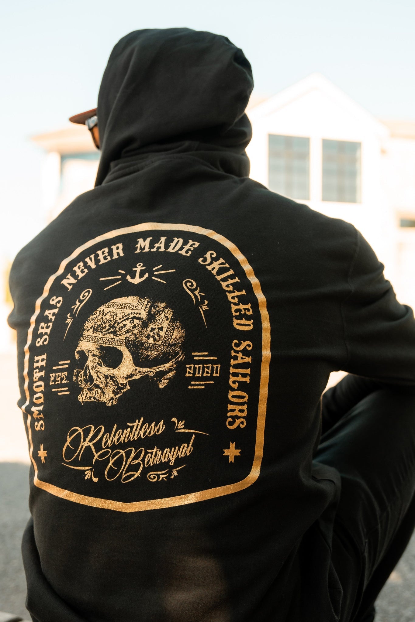 Wearing the Skilled Sailor Gold Foil Hoodie from Relentless Betrayal, made of a cozy cotton/polyester blend, a person relaxes outdoors. The unisex design shows a skull with nautical symbols and Smooth seas never made skilled sailors on the back.