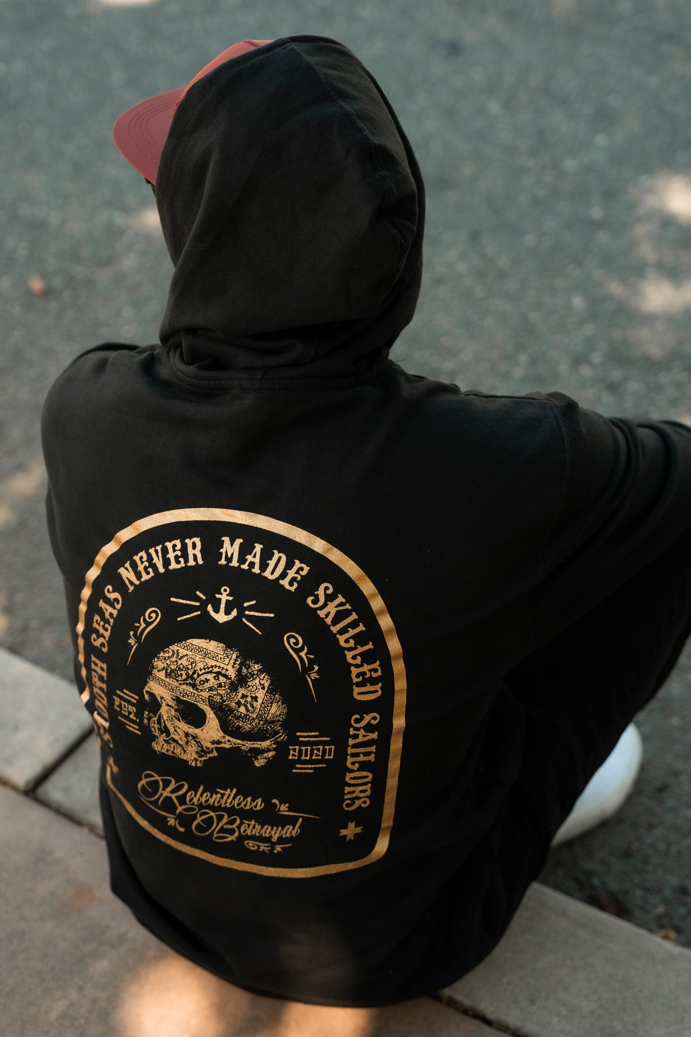 A person sits on a curb wearing a Skilled Sailor Gold Foil Hoodie from Relentless Betrayal in black, featuring a gold anchor and ship wheel design with the phrase Calm Seas Never Made Skilled Sailors. A red cap worn backward completes the unisex look on the gravel-covered ground.