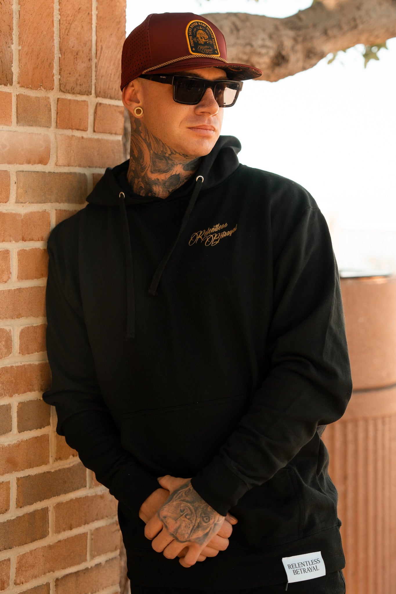 A man poses against a brick wall in a Skilled Sailor Gold Foil Hoodie by Relentless Betrayal, crafted from a cotton/polyester blend. He wears sunglasses and a maroon cap, displaying tattoos on his neck and hands with crossed arms. A tree is visible behind him.