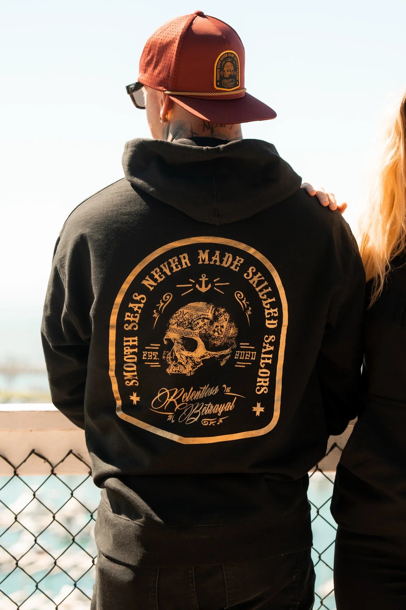 Wearing a Skilled Sailor Gold Foil Hoodie by Relentless Betrayal, made from a comfy cotton/polyester blend, someone stands with their back turned. The black design features a skull, anchor, and the phrase Smooth seas never made skilled sailors in gold foil. Theyre wearing a red cap against the blurred seascape backdrop.