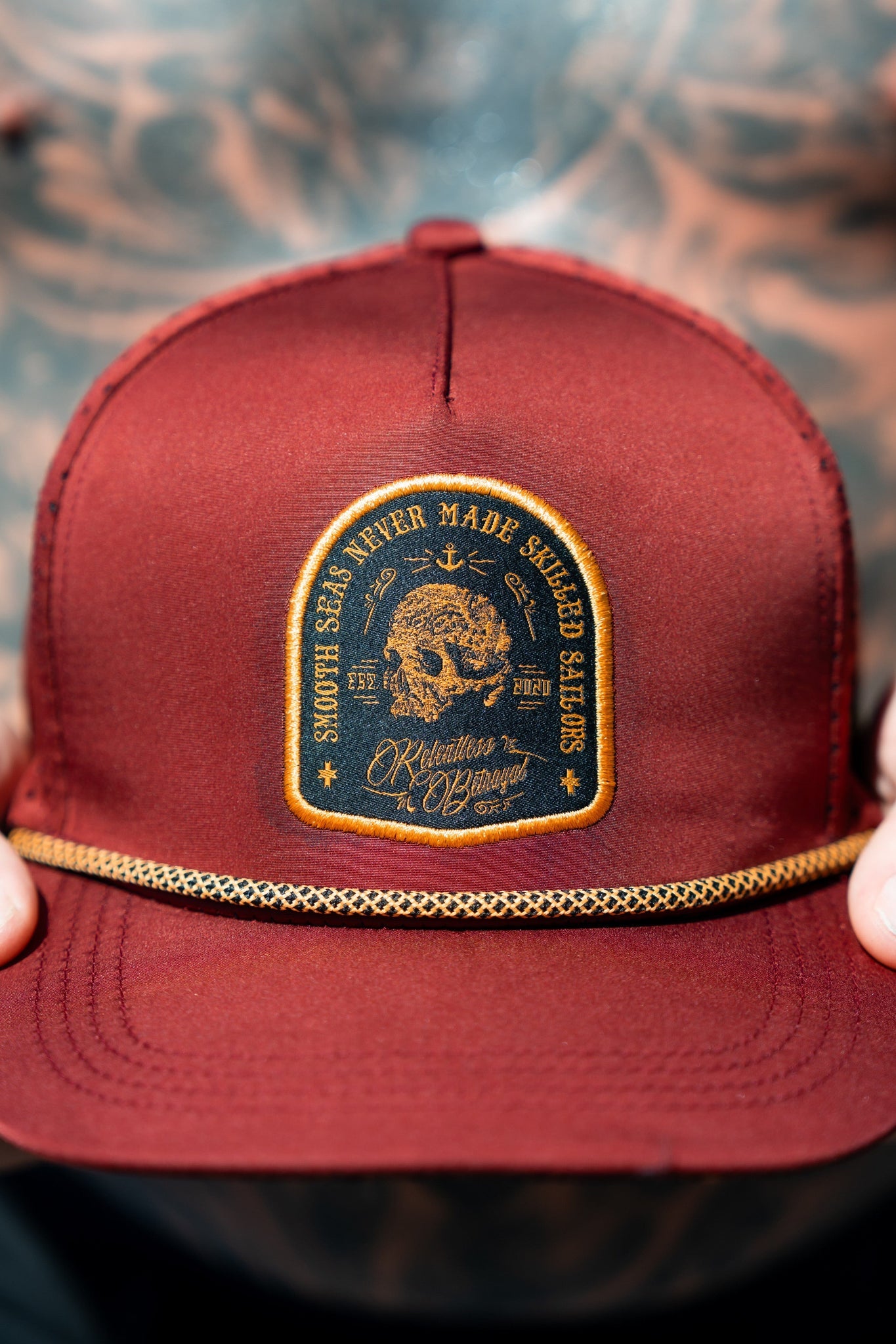 A tattooed person proudly holds a handmade Skilled Sailor Snapback - Burgundy by Relentless Betrayal. The hat features an embroidered ship patch with the text Smooth Seas Never Made Skilled Sailors and Killative x KBangz, complemented by a rope-like trim above the brim.