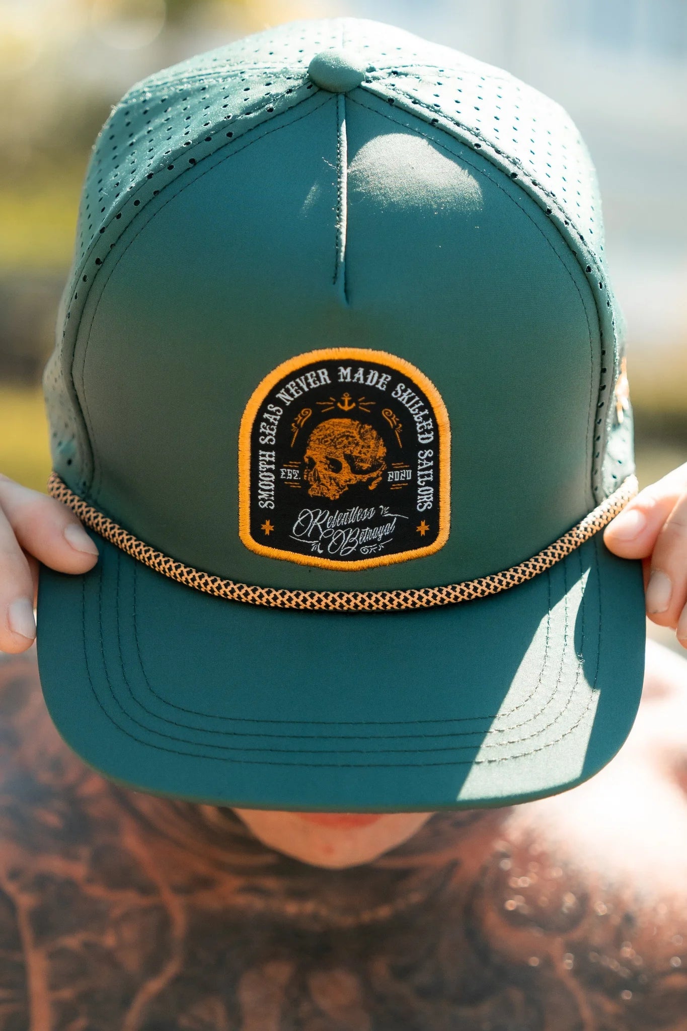 A person dons the Skilled Sailor Snapback in green from Relentless Betrayal, featuring a mesh back and snapback enclosure. They hold the brim adorned with an embroidered patch reading Smooth seas never made skilled sailors and Relentless Pursuit, capturing a sailors spirit.