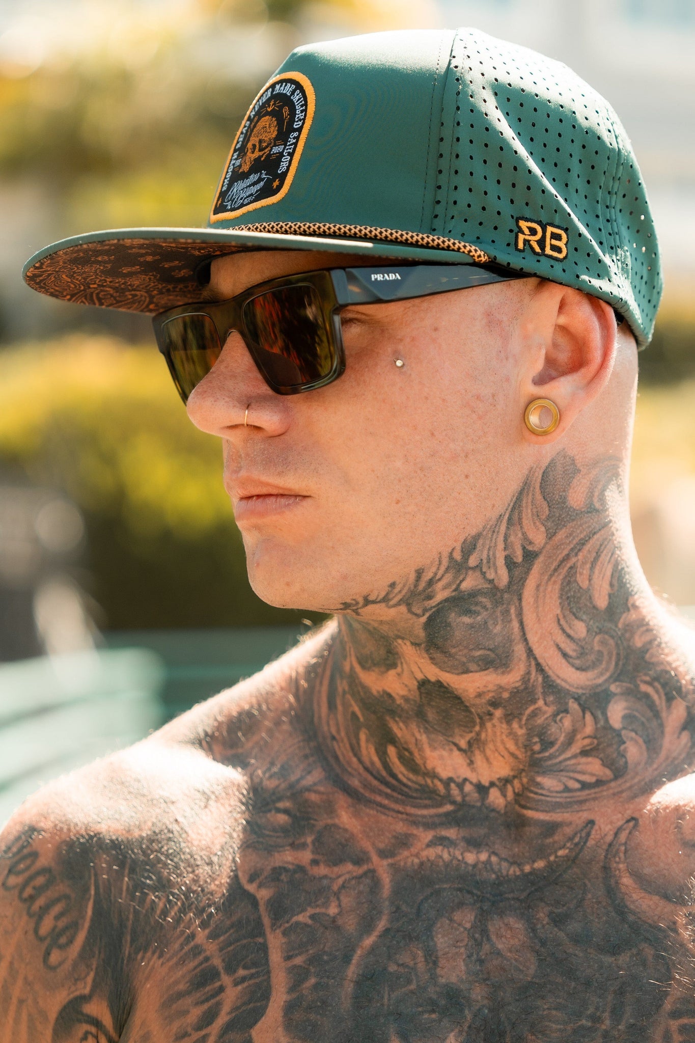 A tattooed individual with ear and nose piercings wears sunglasses and the Skilled Sailor Snapback-Green by Relentless Betrayal, which features an RB embroidery. The snapback closure ensures a snug fit as they stand shirtless against a background of blurred greenery.