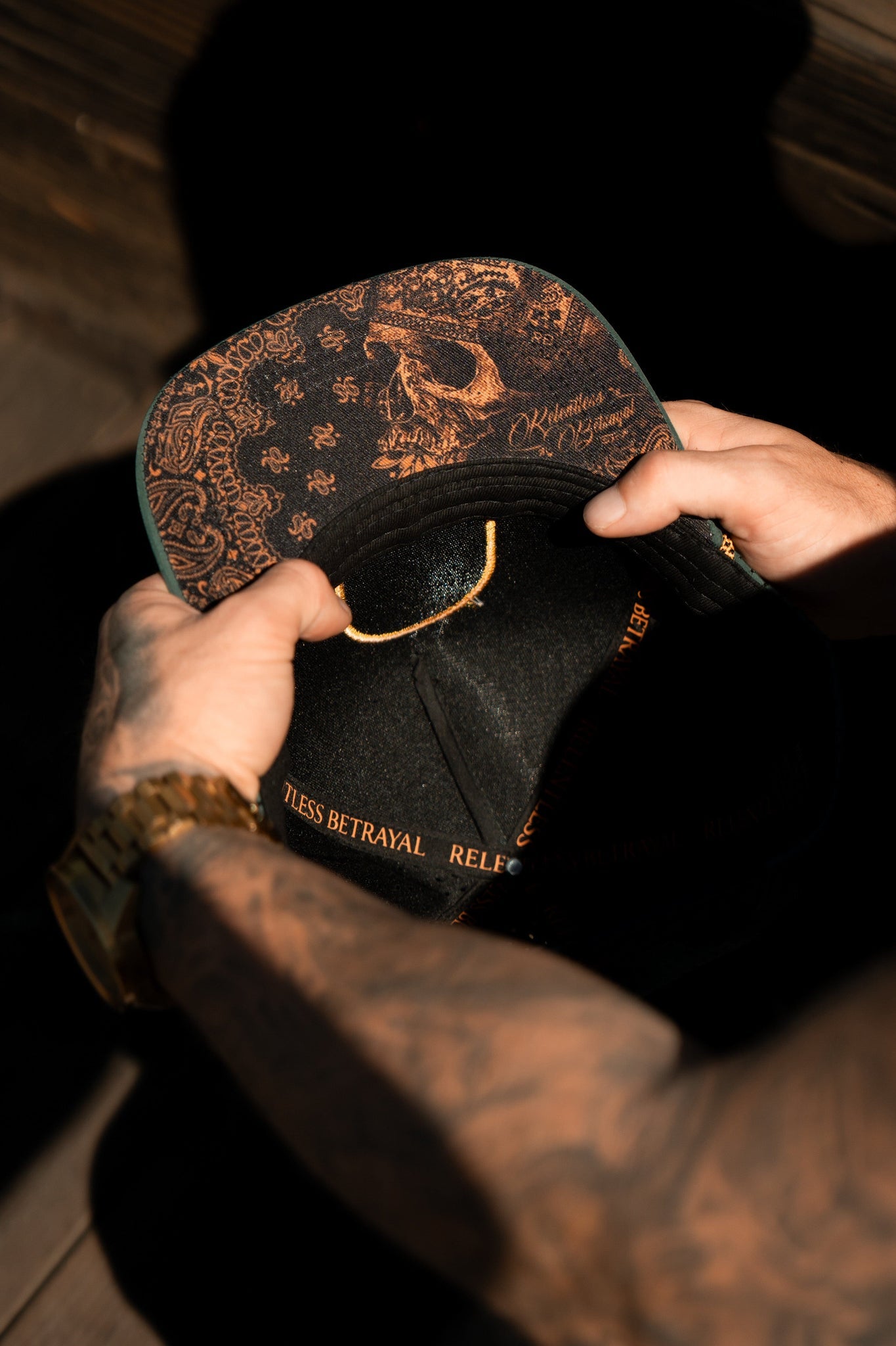 A tattooed individual resembling a sailor is holding a Skilled Sailor Snapback in green by Relentless Betrayal. The underside of the brim has intricate designs and text, with their watch and the textured snapback enclosure visible.