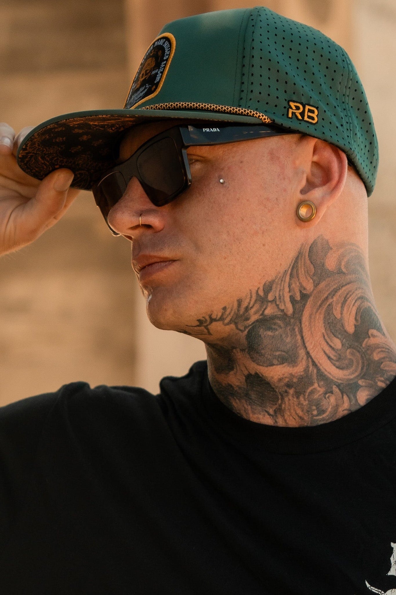 A person with a tattooed neck wears a green Skilled Sailor Snapback by Relentless Betrayal, featuring a snapback enclosure and gold initials RB, dark sunglasses, and a black shirt. They have earrings and a facial piercing outdoors.