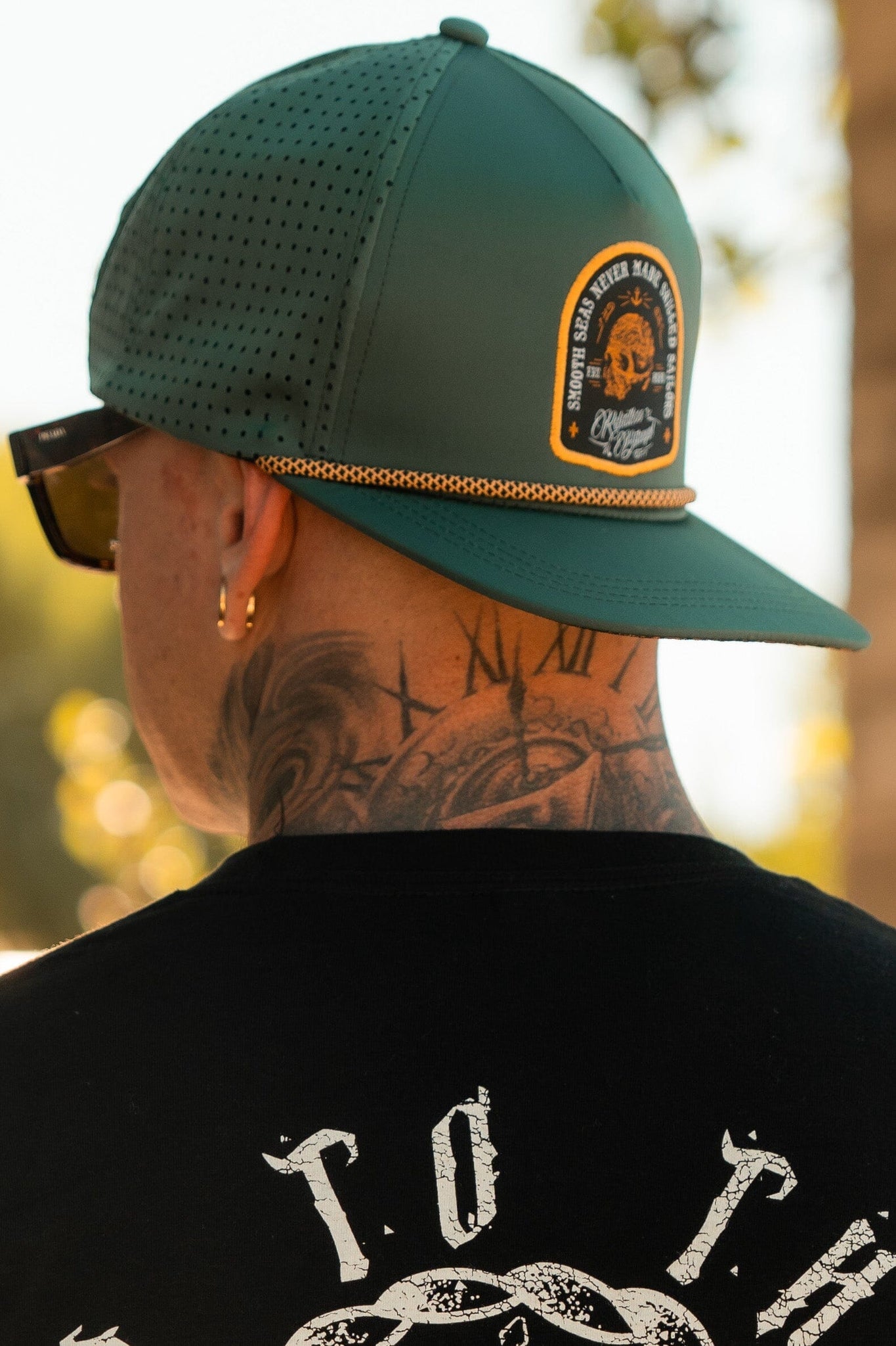 A tattooed person with gold earrings wears Relentless Betrayals Skilled Sailor Snapback in green, featuring a lion patch and snapback closure. Sunglasses cover their eyes, paired with a black shirt sporting bold white lettering on the back.