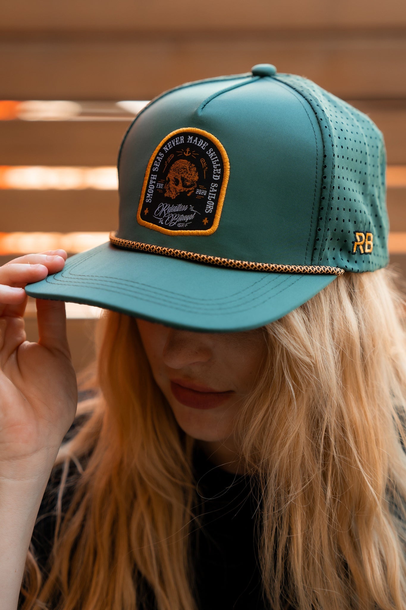 A person with long blond hair wears the Skilled Sailor Snapback- Green by Relentless Betrayal. This water-resistant hat has perforated sides, a snapback enclosure, and a black patch with deer and tree graphics, featuring the text Shredded & Never Made Stag Slabs BiltHammer Design.