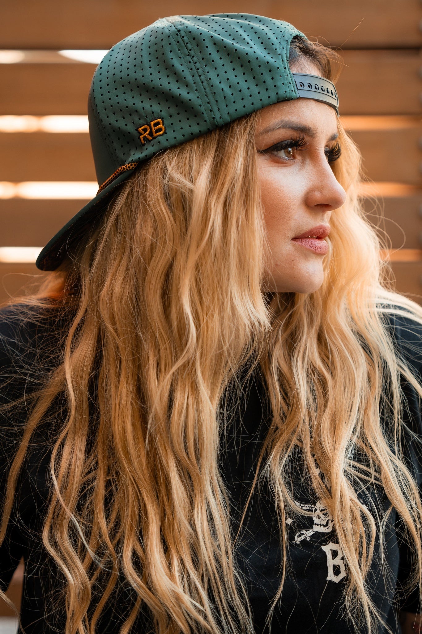 A person with long, wavy blonde hair wears a Skilled Sailor Snapback in green by Relentless Betrayal backwards, paired with a black shirt featuring a white logo. They glance to the side against wooden slats, embodying effortless style.