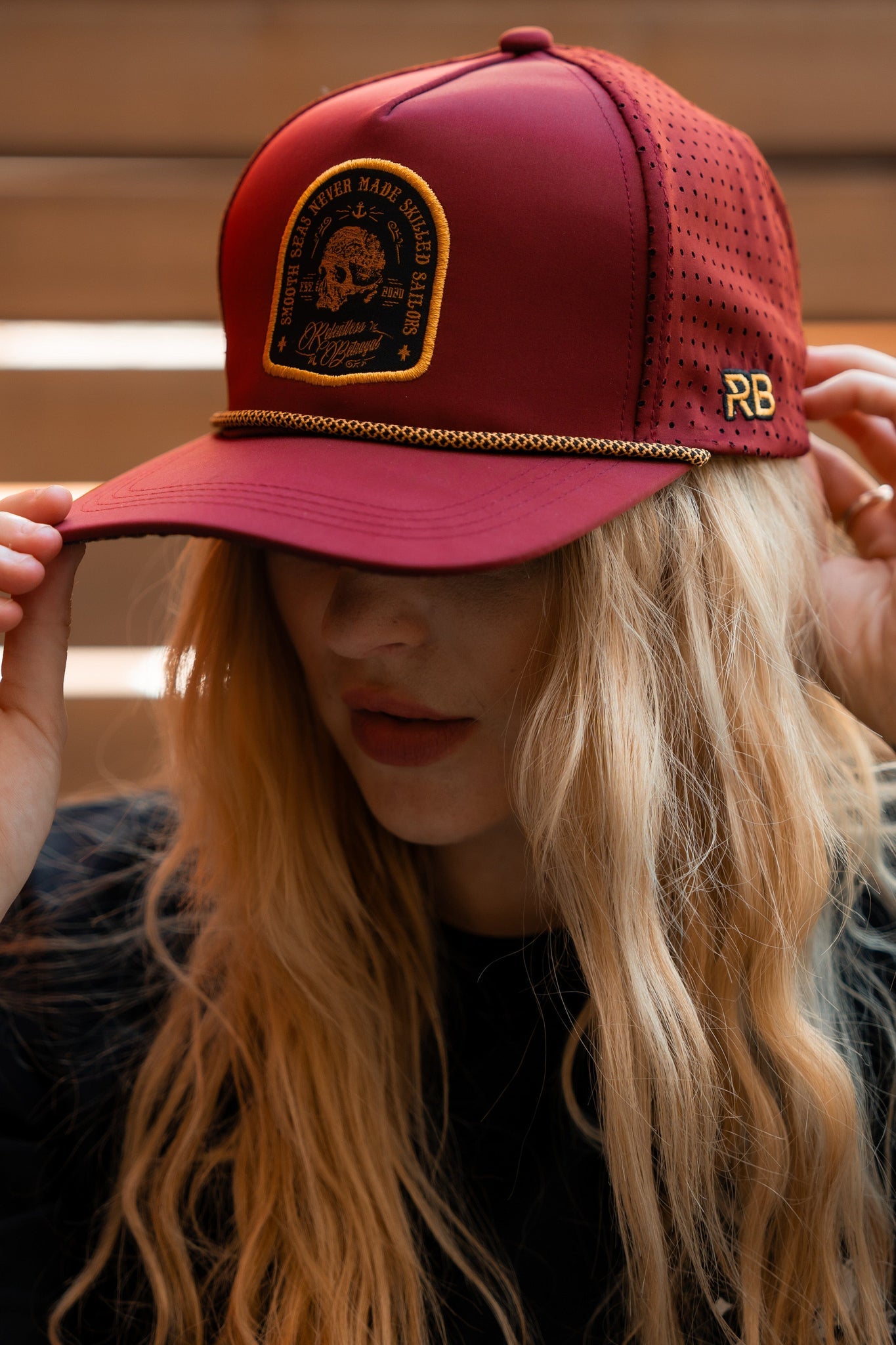 A person with long blonde hair adjusts the Relentless Betrayal Skilled Sailor Snapback in burgundy, adorned with gold chain detail, black text, and a graphic. It stands out against a blurred wooden background with its unique flair.
