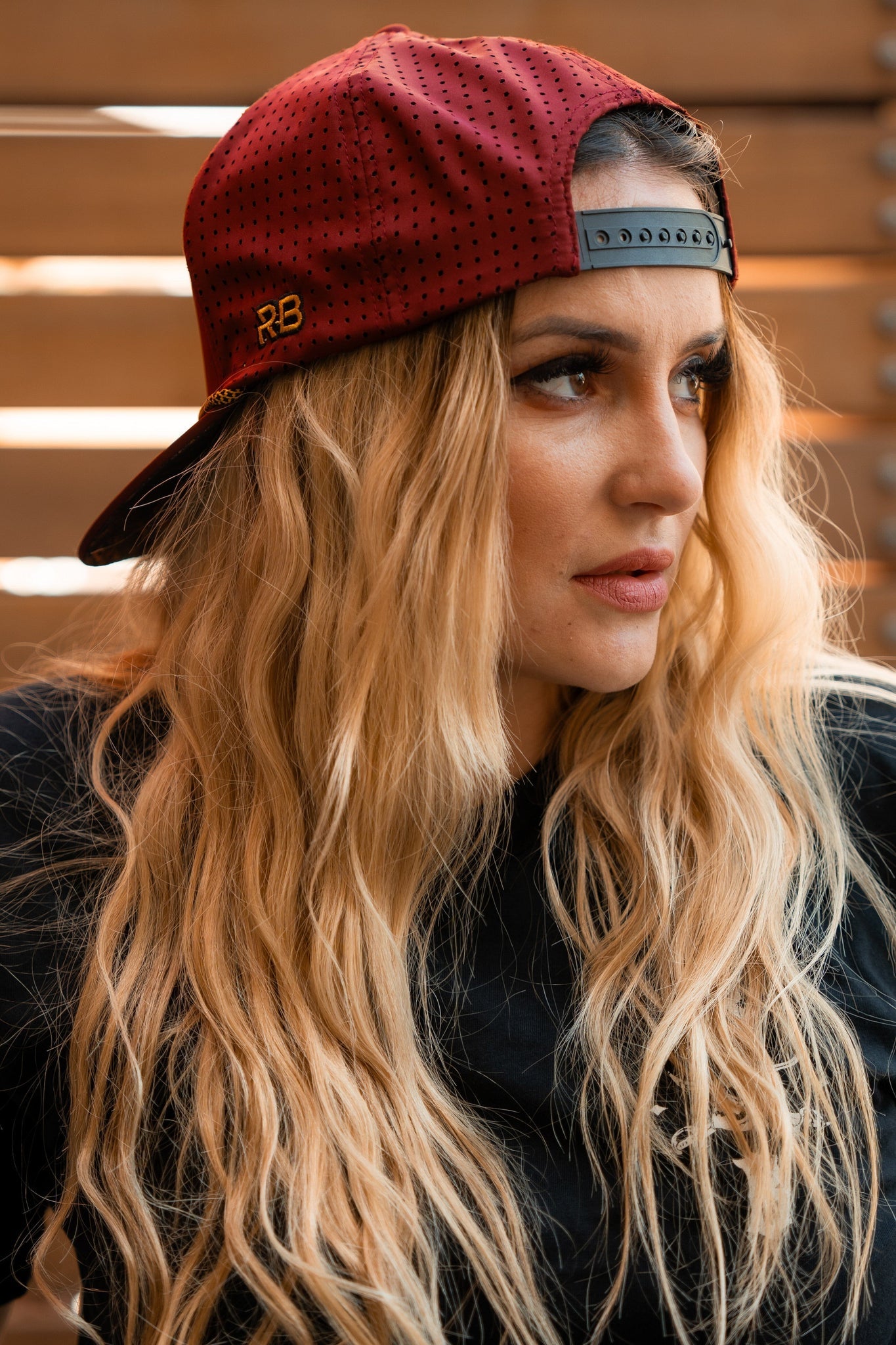 A person with long, wavy blonde hair wears a Skilled Sailor Snapback - Burgundy from Relentless Betrayal backward and a black shirt, looking to the side. The warmly lit background features horizontal wooden planks.