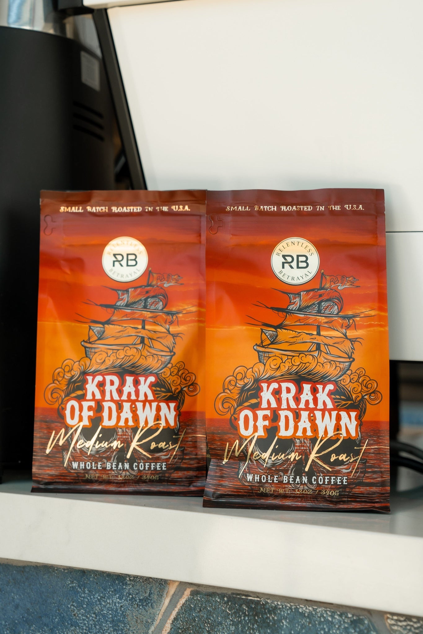 Two bags of Relentless Betrayals Krak Of Dawn medium roast coffee, crafted from Colombian Arabica beans, sport packaging with a ship on an orange background, emphasizing its small batch roasting in the USA.