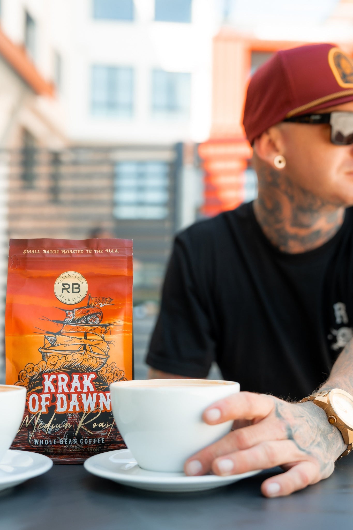 A tattooed person in a red cap and sunglasses sits at a table with two white cups, displaying a bag of Relentless Betrayals Krak Of Dawn medium roast coffee, made from Rainforest Alliance Certified Colombian Arabica beans. The background showcases a modern building with large windows.