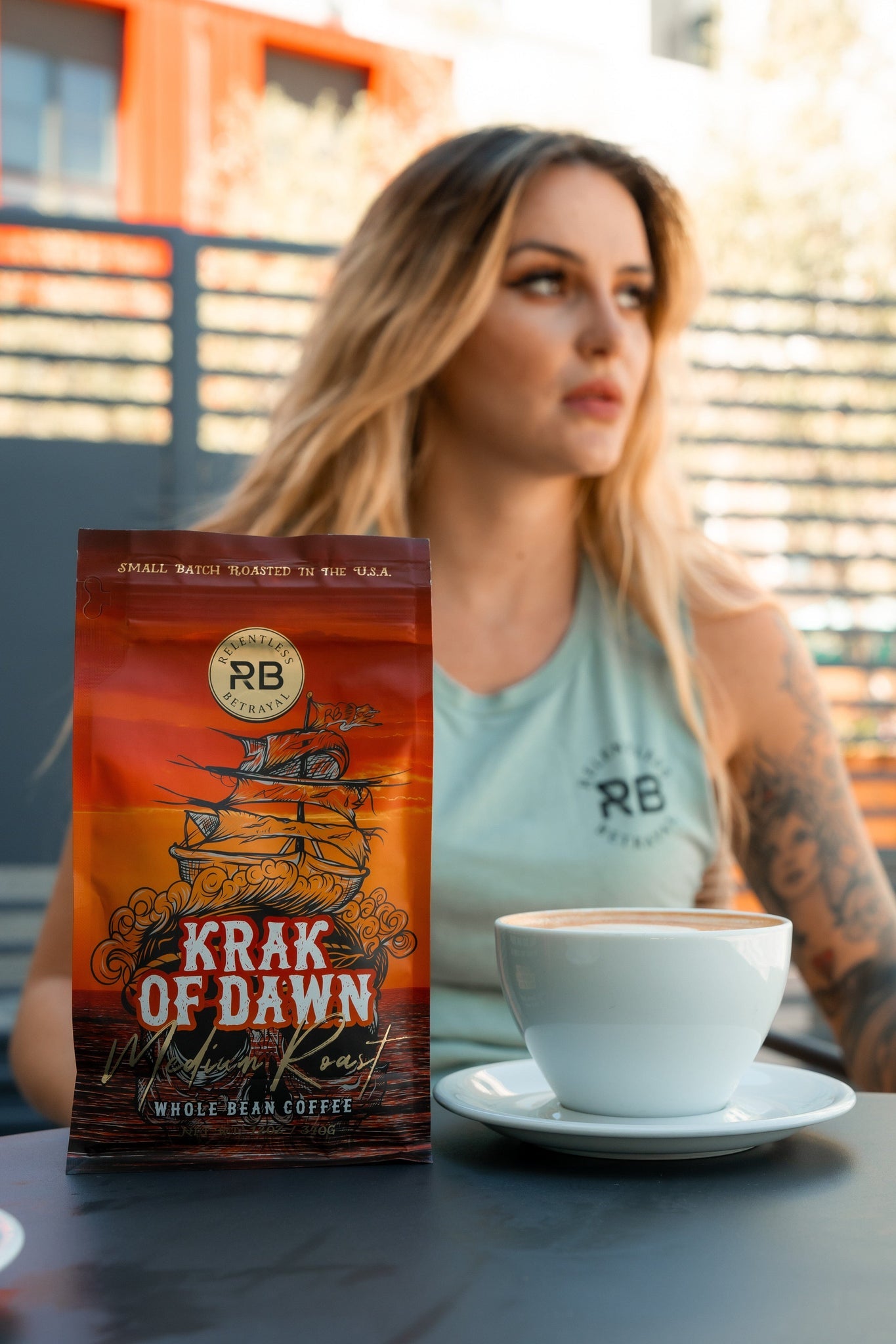 A woman with long hair and tattoos sits at a table, wearing a light green sleeveless top. In front of her is a cup of coffee next to Krak Of Dawn Coffee by Relentless Betrayal, made from Rainforest Alliance Certified Colombian Arabica beans. The scene is brightly lit.