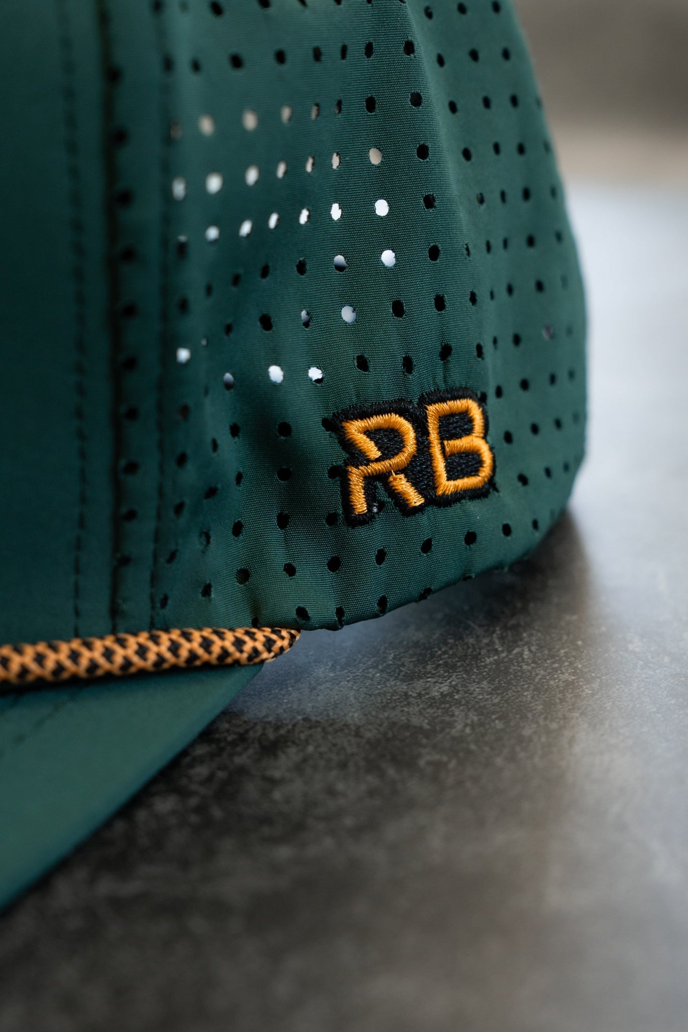 Close-up of the Skilled Sailor Snapback – Green by Relentless Betrayal, featuring orange RB embroidery, small perforations, an orange stitched border along the edge, and a snapback enclosure for a perfect fit.