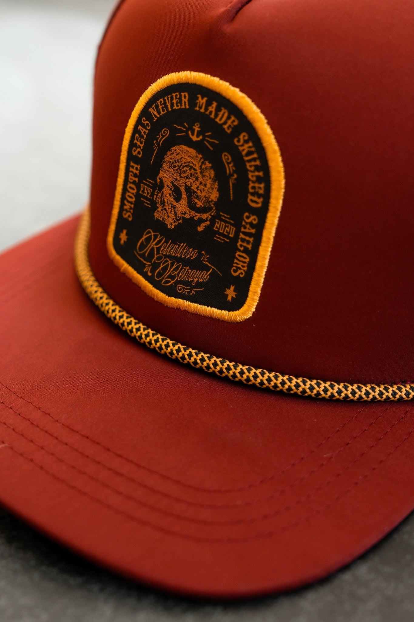 The Relentless Betrayal Skilled Sailor Snapback - Burgundy is a water-resistant cap featuring yellow embroidered nautical symbols and the phrase Smooth Seas Never Made Skilled Sailors on a maroon background, with a decorative yellow rope above the brim.