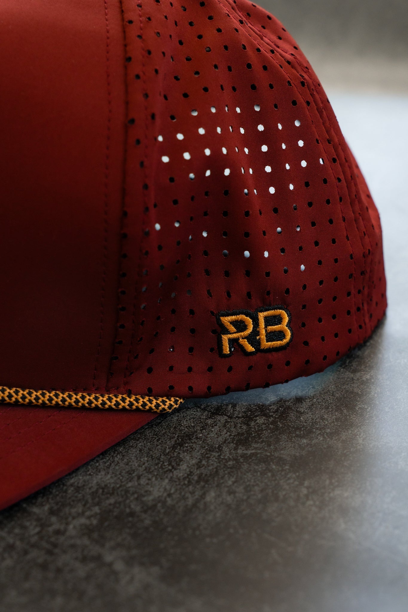 Close-up of the Skilled Sailor Snapback - Burgundy by Relentless Betrayal, featuring perforated fabric with black dots, orange stitching, and yellow RB initials. This water-resistant snapback lies on a gray surface, highlighting its sleek and stylish design.