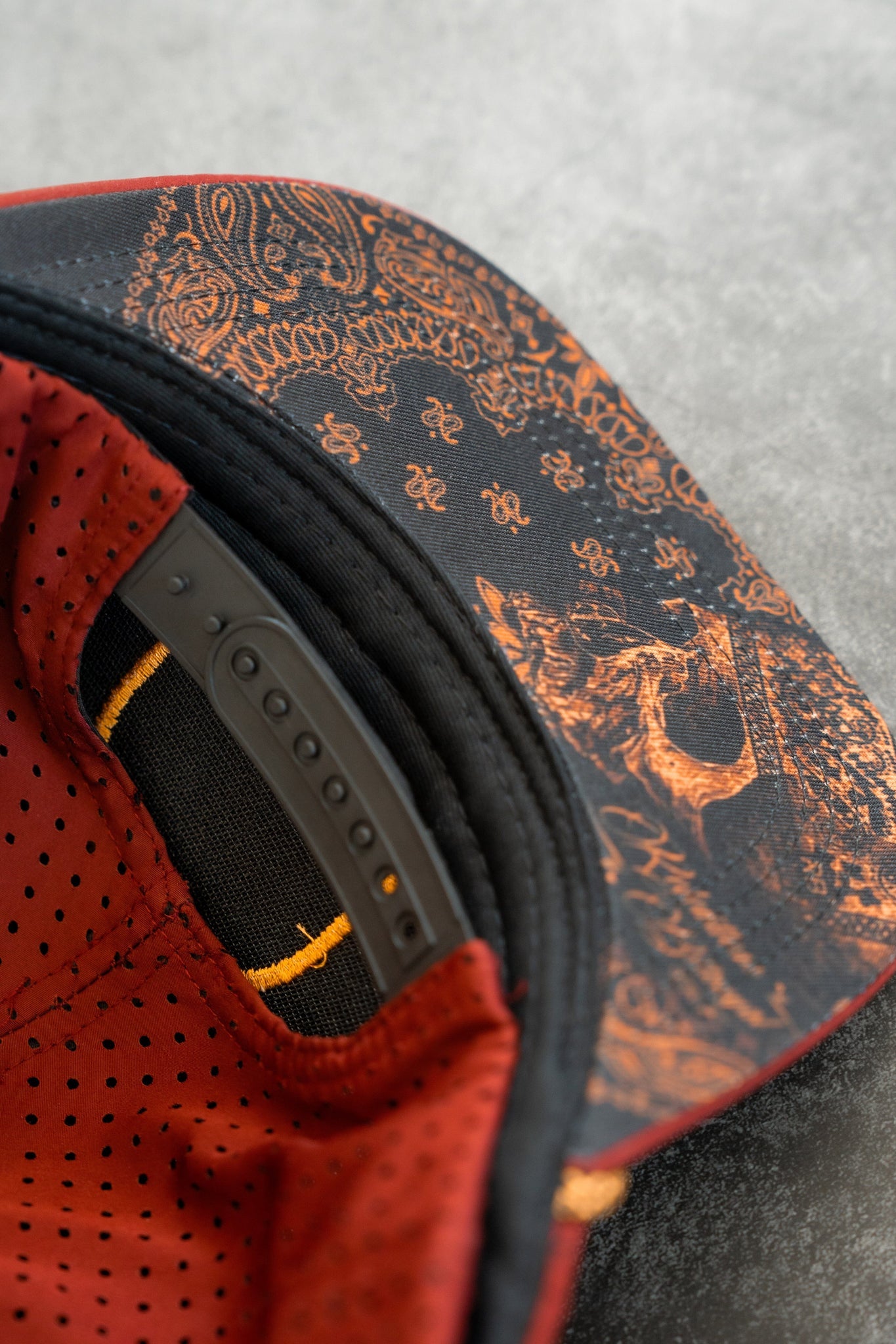 Close-up of a water-resistant Relentless Betrayal snapback featuring ornate orange patterns and a bear under the brim. Inside, it has a black adjustable strap and red perforated fabric. The textured gray backdrop highlights this Skilled Sailor Snapback - Burgundy craftsmanship.