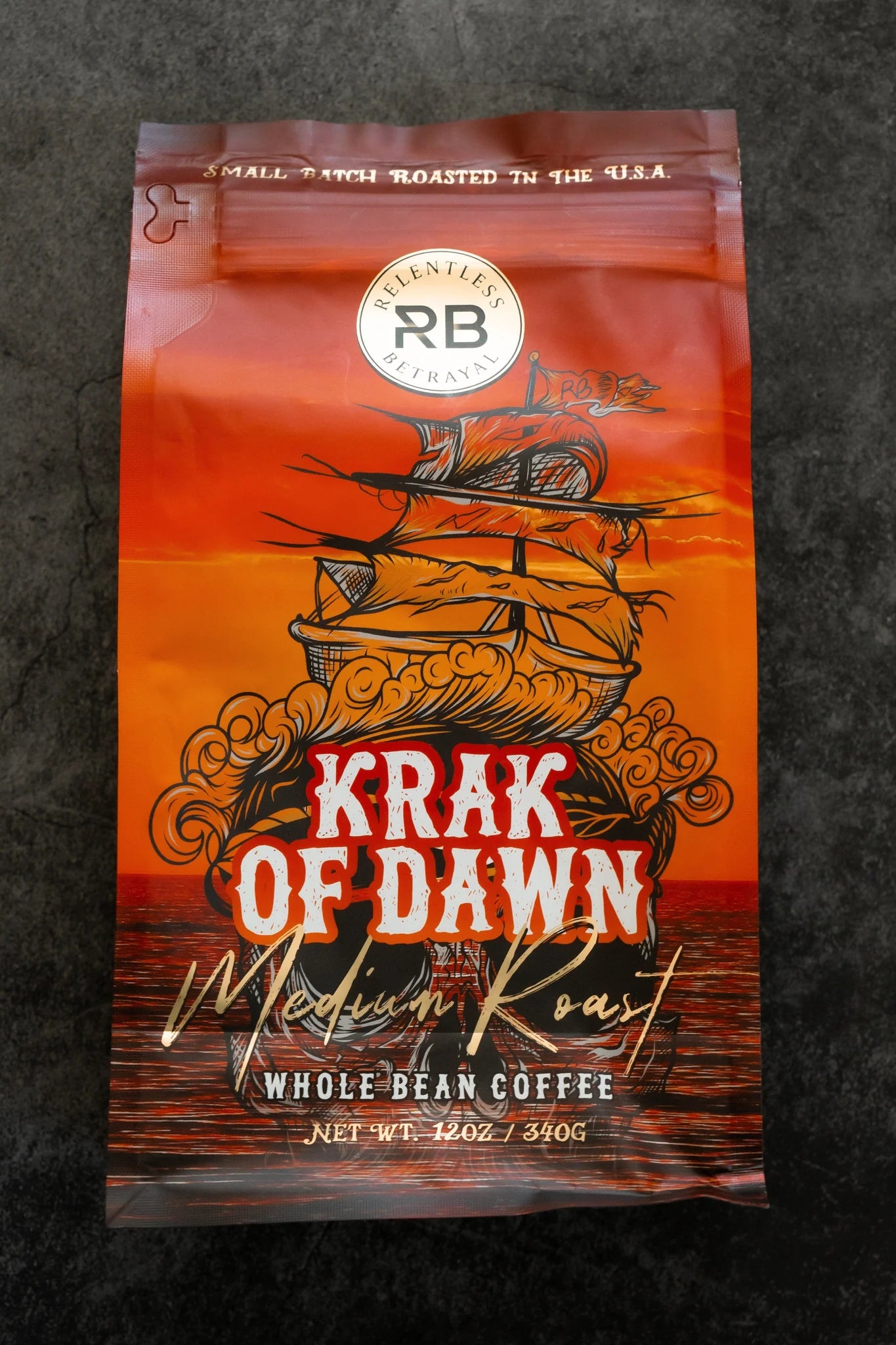A bag of Krak Of Dawn Coffee from Relentless Betrayal sits on a dark surface. The design features a vintage ship against an orange background with swirling clouds, highlighting its Rainforest Alliance Certified whole bean coffee status, net weight 12 oz.