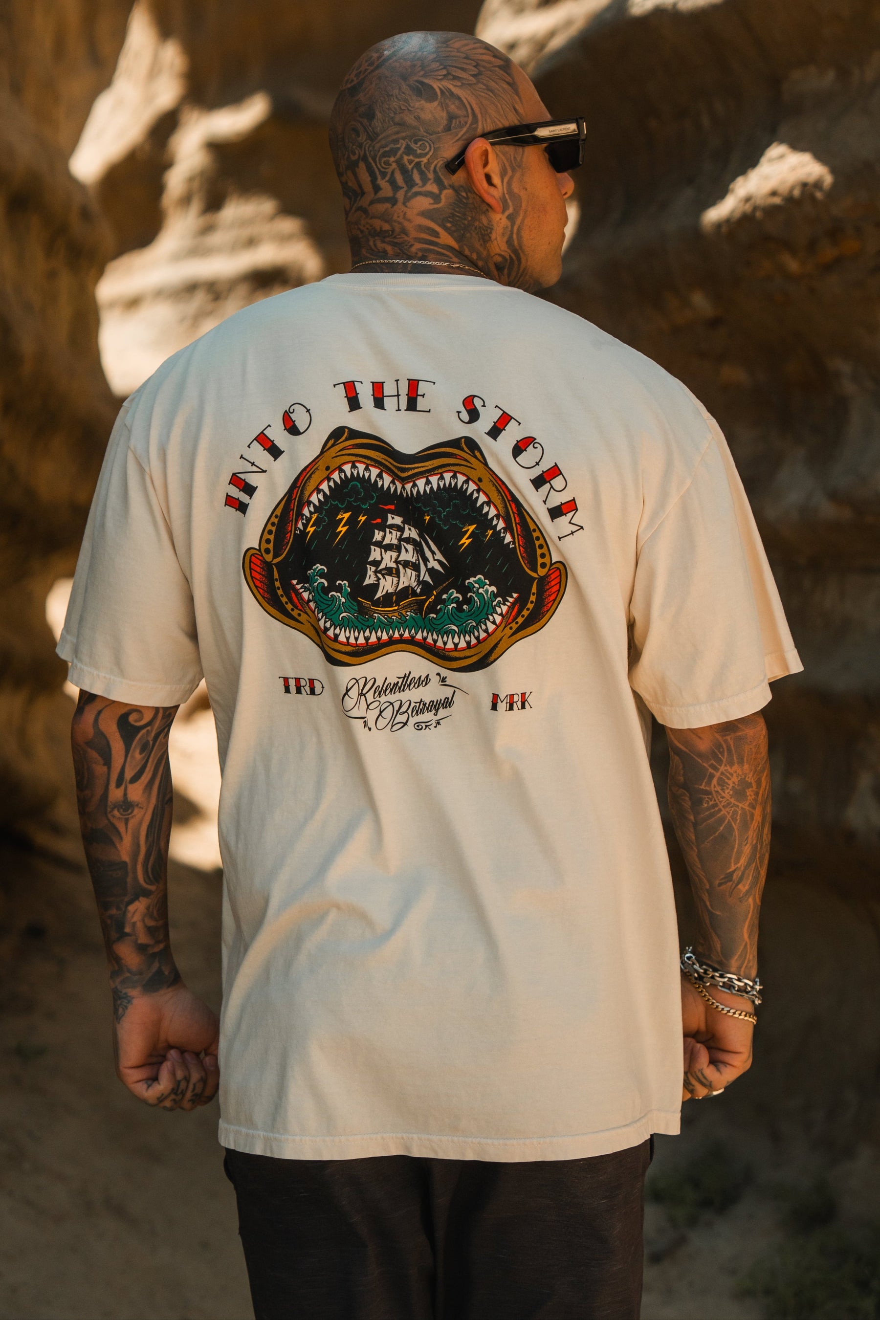 A tattooed person in sunglasses wears the Relentless Betrayal Into the Storm Premium Ivory T-Shirt, featuring a large ship graphic and “INTO THE STORM” on premium jersey cotton, standing outdoors with their back to the camera.
