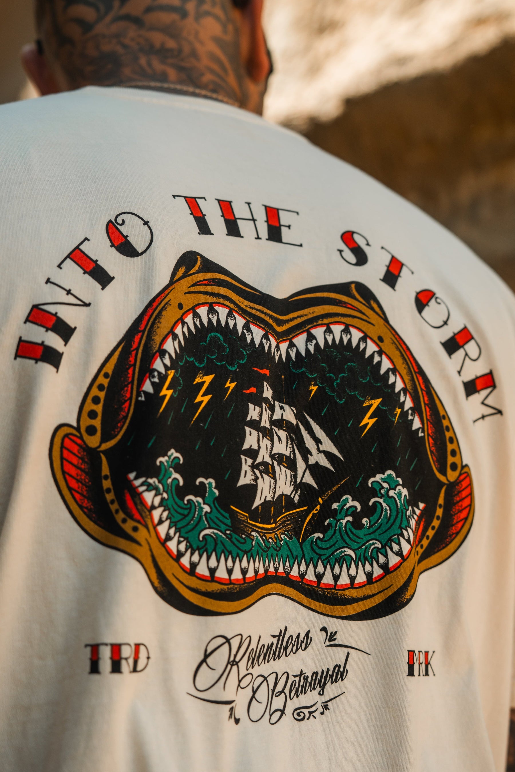 A person wears the Relentless Betrayal Into the Storm Premium Ivory T-Shirt, featuring a graphic of an open-mouthed sea monster, ship, waves, lightning, and INTO THE STORM text. Made from premium jersey cotton.