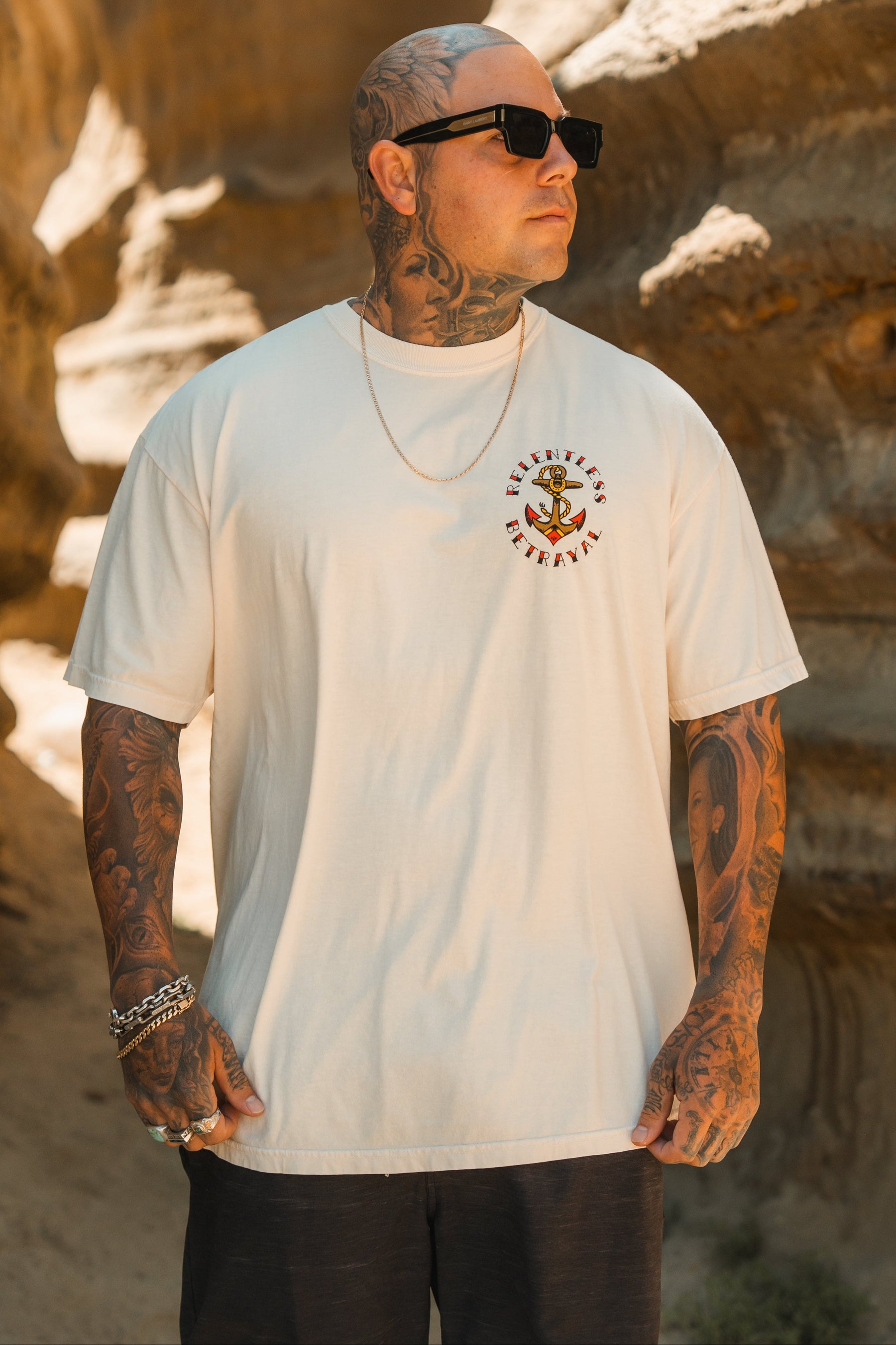 A tattooed man in sunglasses and a gold chain wears the Relentless Betrayal Into the Storm Premium Ivory T-Shirt, featuring a skull graphic and bold text, as he stands outdoors by rocky formations looking to the side.