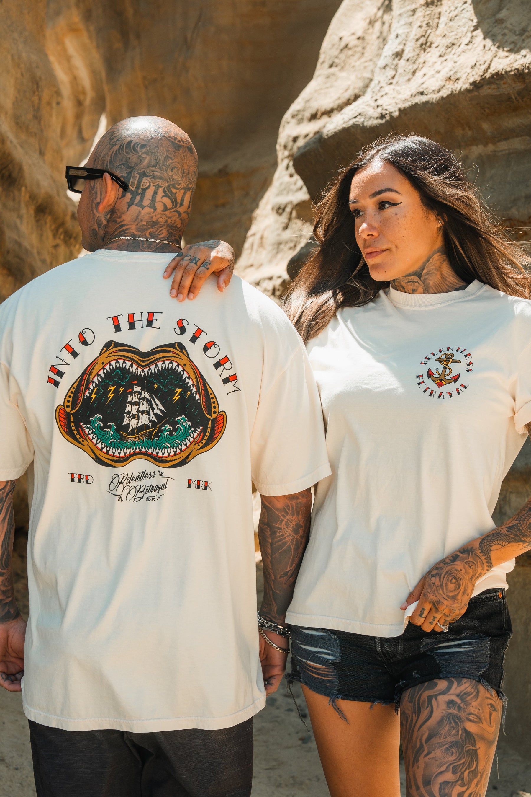 A tattooed man and woman stand near rocks, both in Relentless Betrayal’s Into the Storm Premium Ivory T-Shirts; his features Into the Storm with a ship, while hers displays a small anchor graphic on the front.