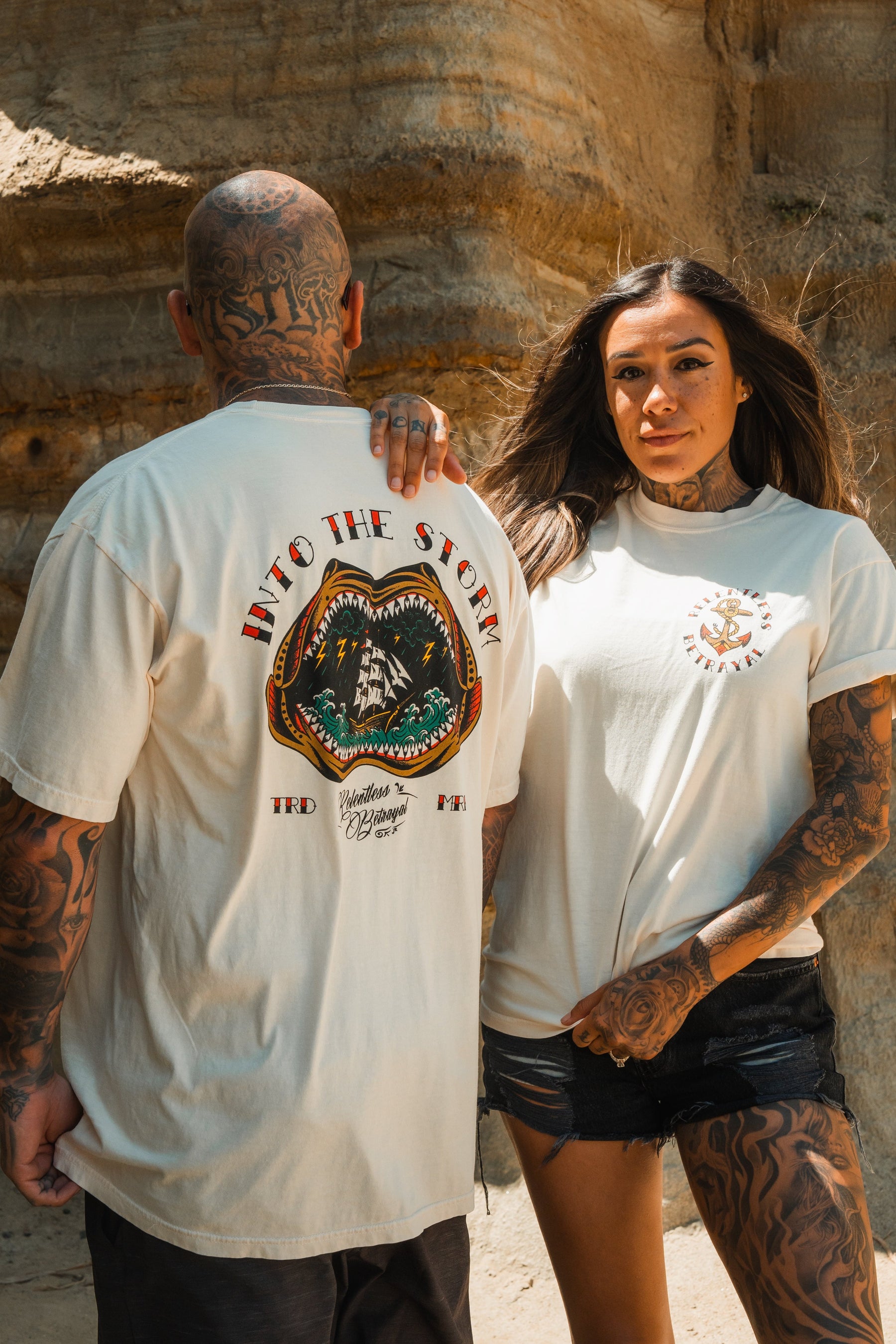 A tattooed man and woman stand by rocky terrain wearing Relentless Betrayal’s Into the Storm Premium Ivory T-Shirt, featuring a mountain and eagle graphic. The woman pairs hers with black shorts.