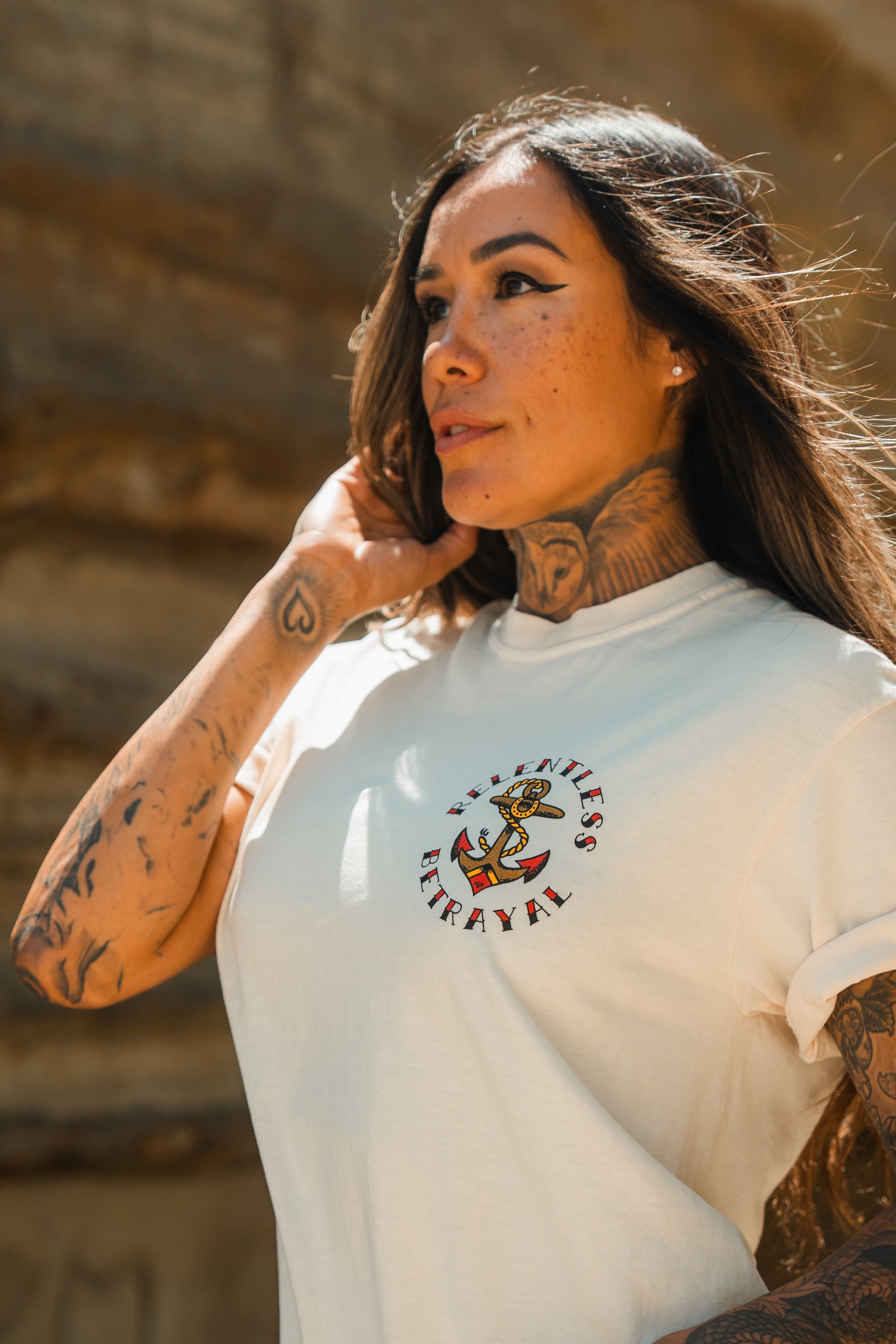 A woman with long hair and tattoos stands outdoors wearing the Relentless Betrayal Into the Storm Premium Ivory T-Shirt, made from jersey cotton and featuring an anchor graphic with the words No Titles, No Betrayal.
