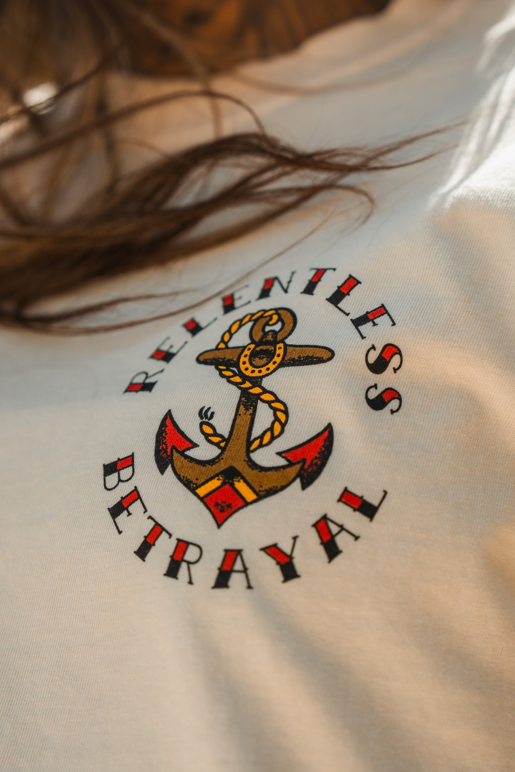 A close-up of the Into the Storm Premium Ivory T-Shirt by Relentless Betrayal, featuring an anchor graphic with red accents and RELENTLESS BETRAYAL in bold around it; brown hair partially frames this premium jersey cotton unisex tee.