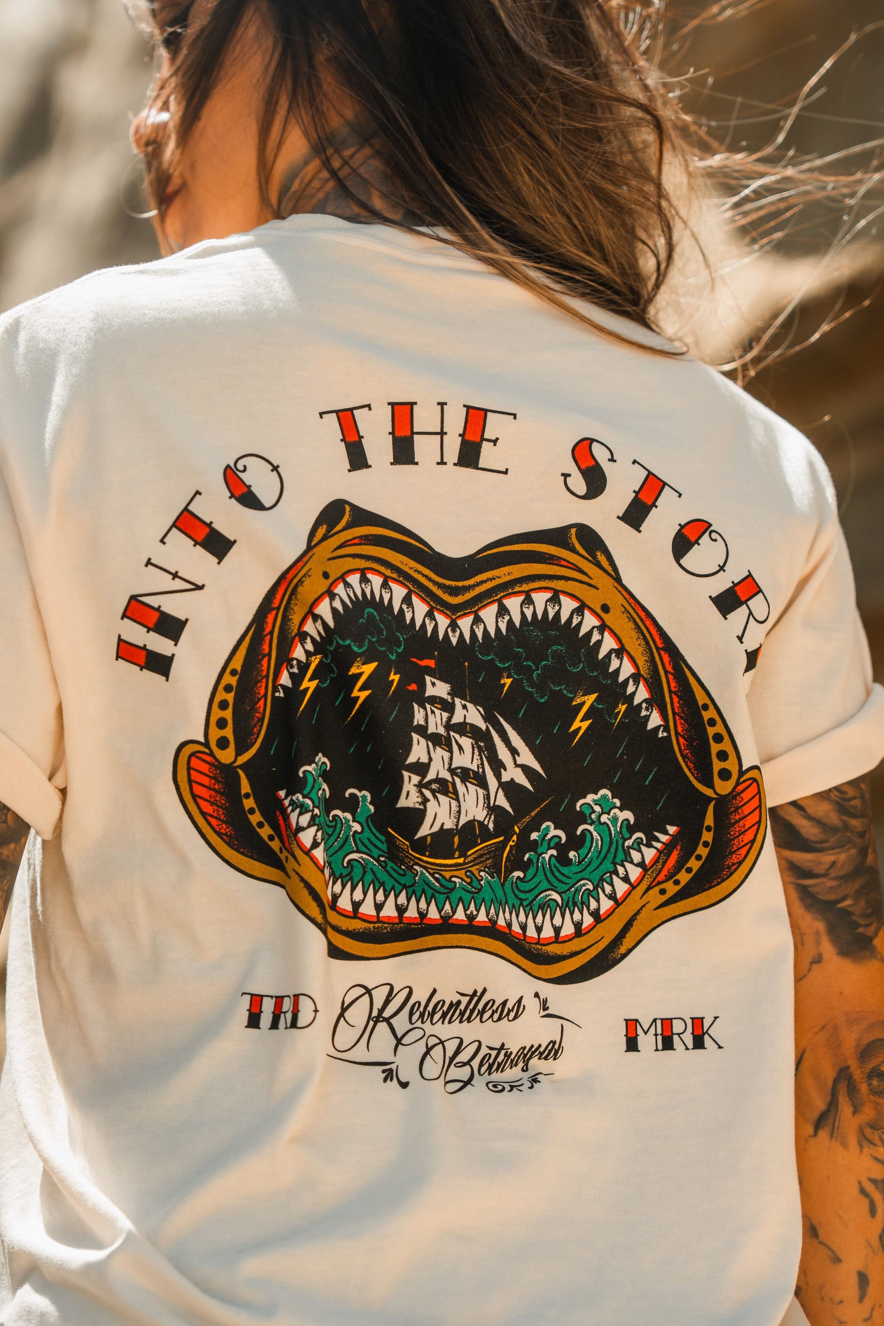Someone wears the Relentless Betrayal Into the Storm Premium Ivory T-Shirt, featuring a vivid back graphic of an open-mouthed sea creature with a ship inside, waves, lightning, and “INTO THE STORM” text above.