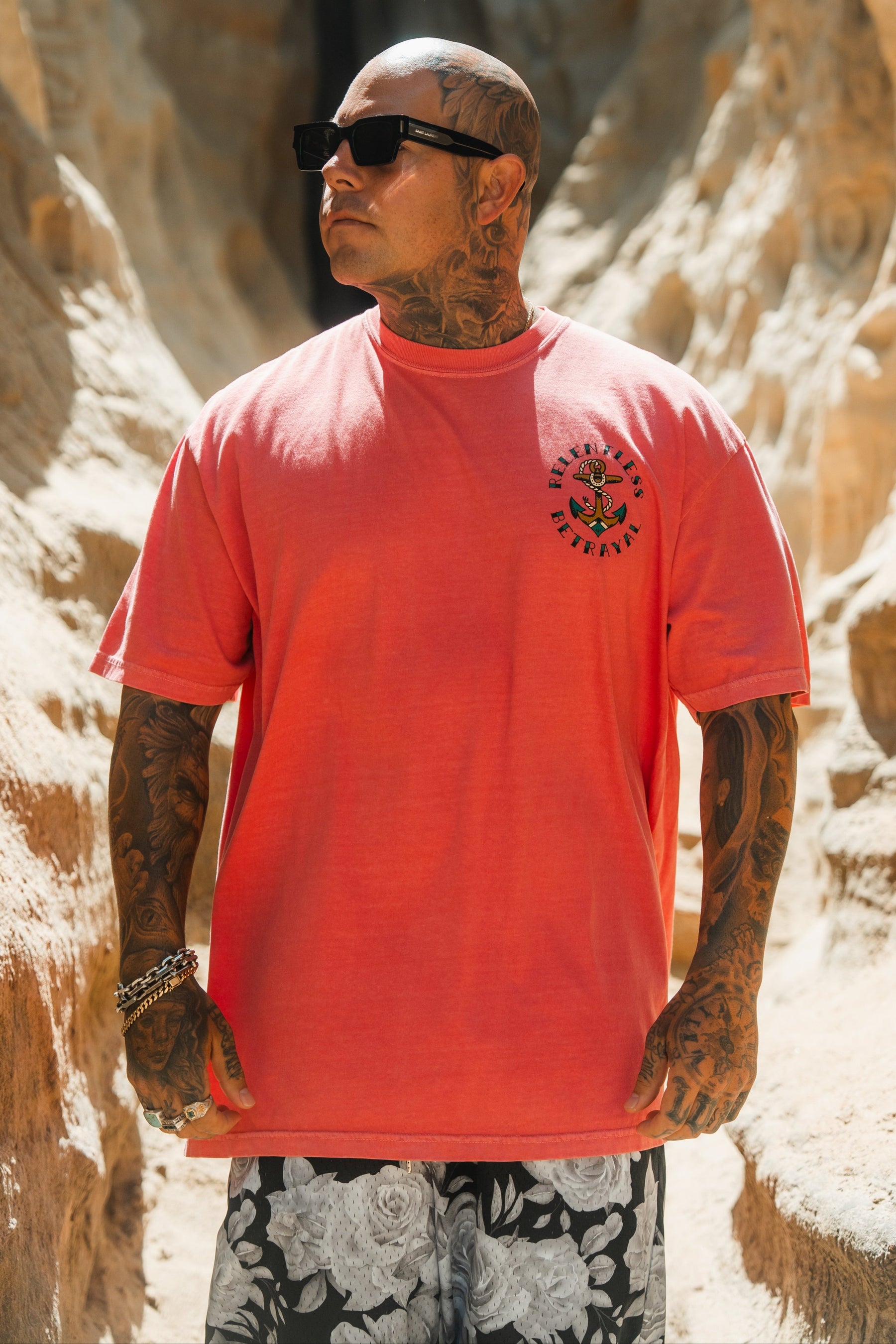A tattooed man in sunglasses stands outdoors among rocks, wearing the Relentless Betrayal Into the Storm Premium Watermelon T-Shirt and black and white floral shorts, sunlight highlighting his face and arms.