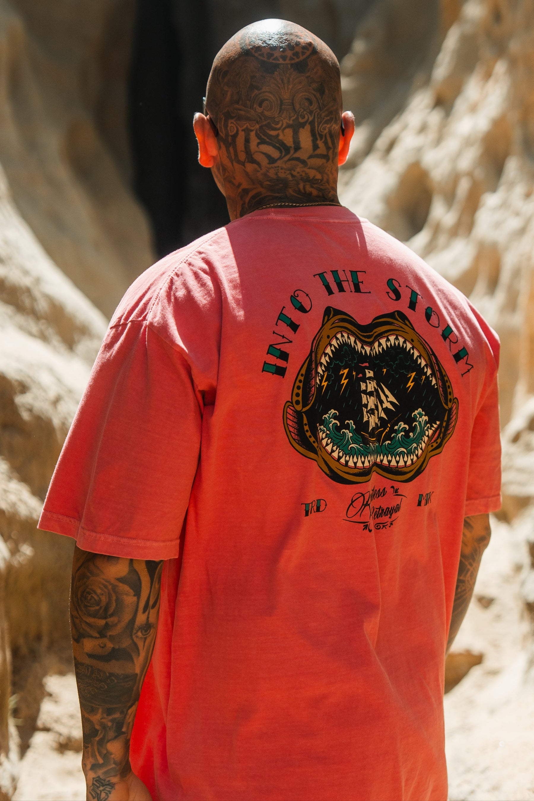 A tattooed person with a shaved head stands near rocky terrain, wearing the Relentless Betrayal Into the Storm Premium Watermelon T-Shirt with a bold ship-in-a-storm graphic and INTO THE STORM text on the back.