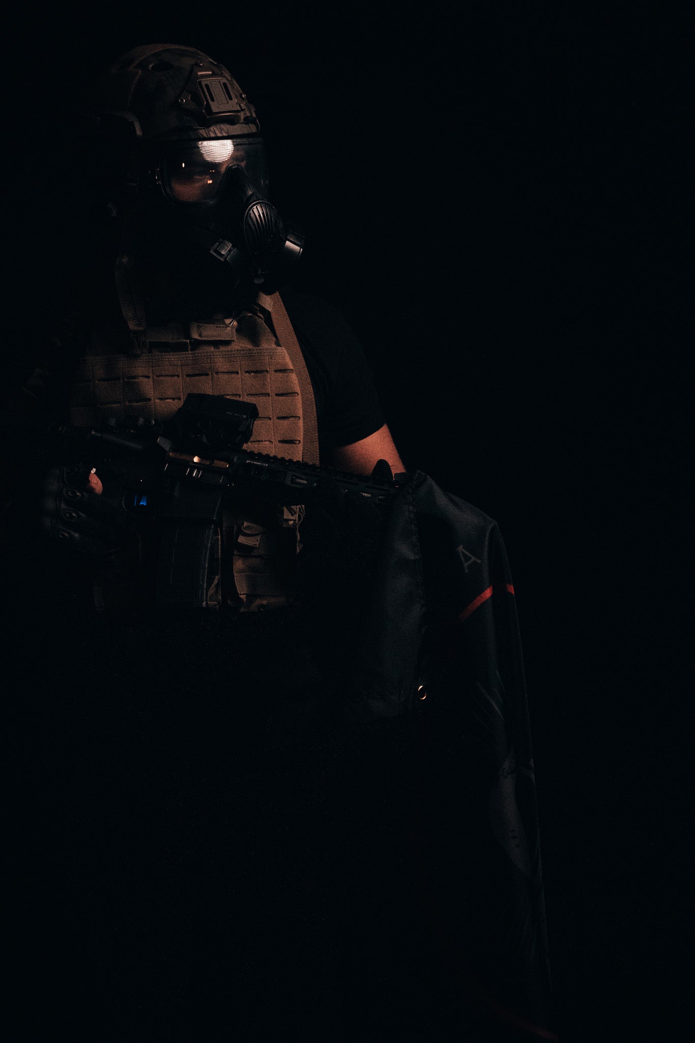 A person wearing tactical gear, including a high-quality helmet and gas mask, stands dramatically in the dark. Their bulletproof vest, reinforced with durable polyester and metal grommets and supporting a rifle, evokes imagery from the Born United Private Labels Saints and Sinners Flag collection.