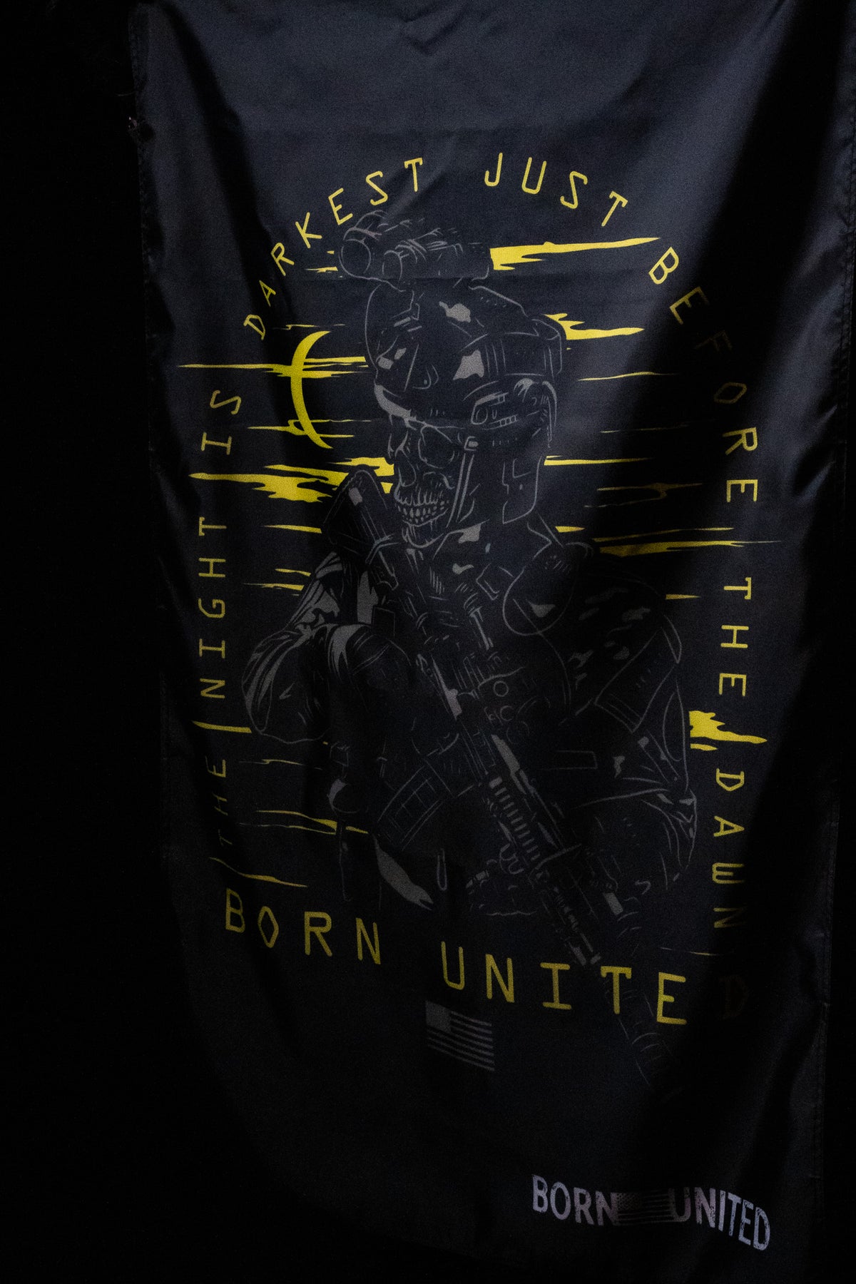 The Before Dawn Flag from Born United Private Label features a polyester design with a skull in a helmet holding a rifle. It includes yellow text, THE NIGHT IS DARKEST JUST BEFORE THE DAWN and BORN UNITED. Metal grommets make hanging easy, while a patch states BORN UNITED.