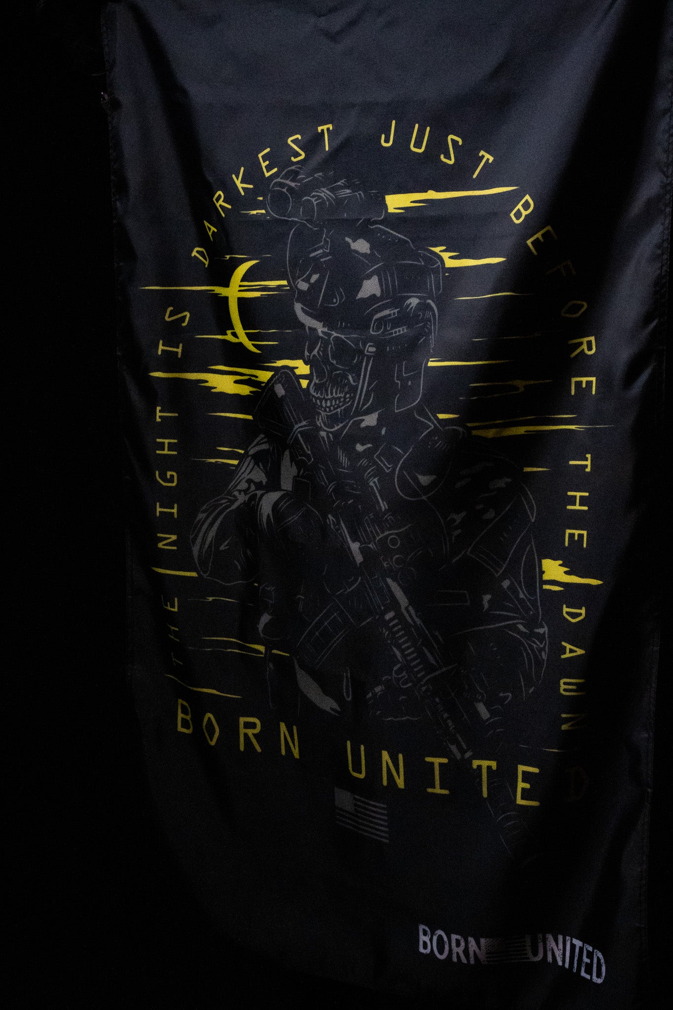 The Before Dawn Flag from Born United Private Label features a polyester design with a skull in a helmet holding a rifle. It includes yellow text, THE NIGHT IS DARKEST JUST BEFORE THE DAWN and BORN UNITED. Metal grommets make hanging easy, while a patch states BORN UNITED.