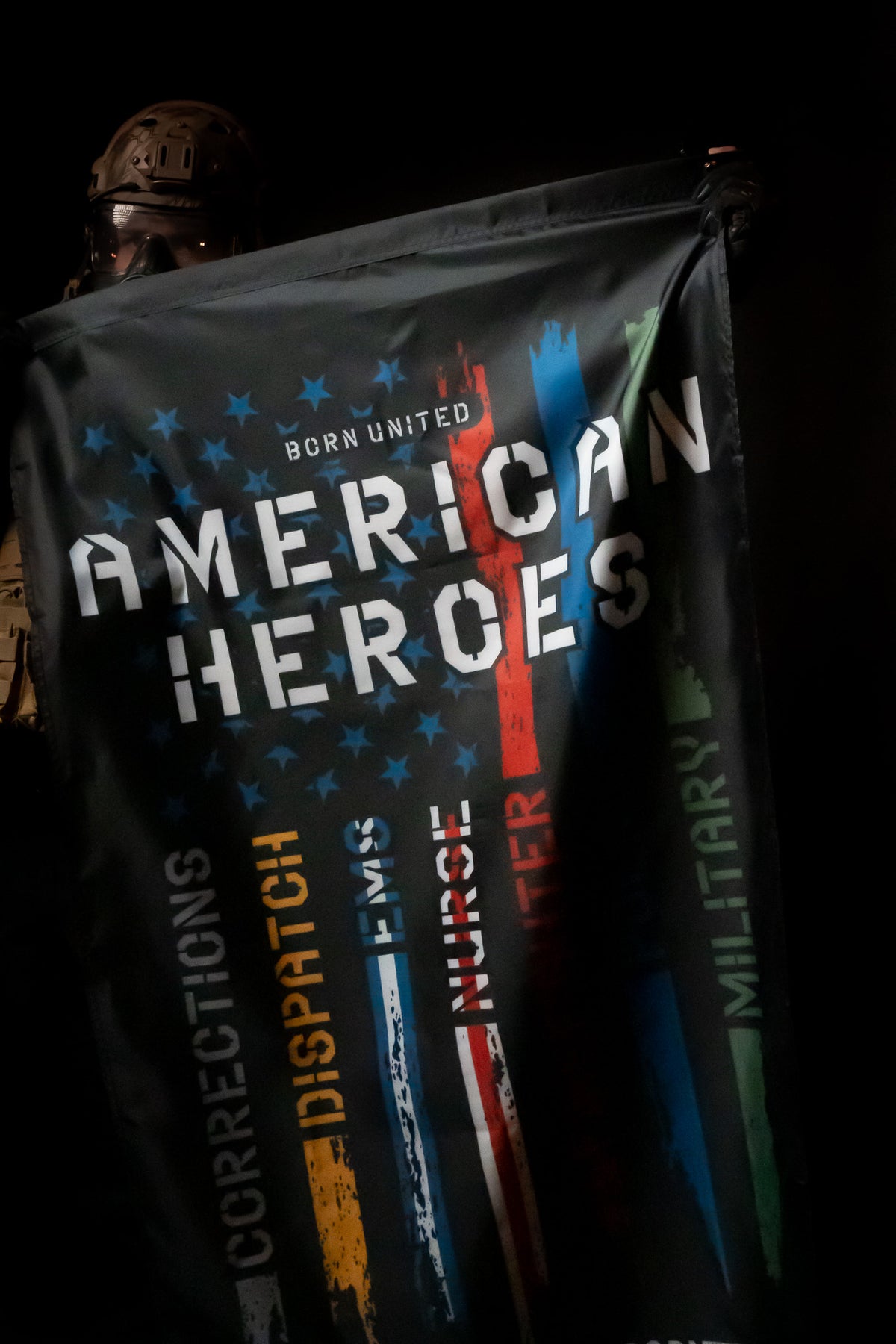 A person in tactical gear holds the American Heroes Flag from Born United Private Label, showcasing vertical stripes to honor first responders and military, featuring American Heroes and words like Corrections, Dispatch, and EMS, all set against stars and stripes.