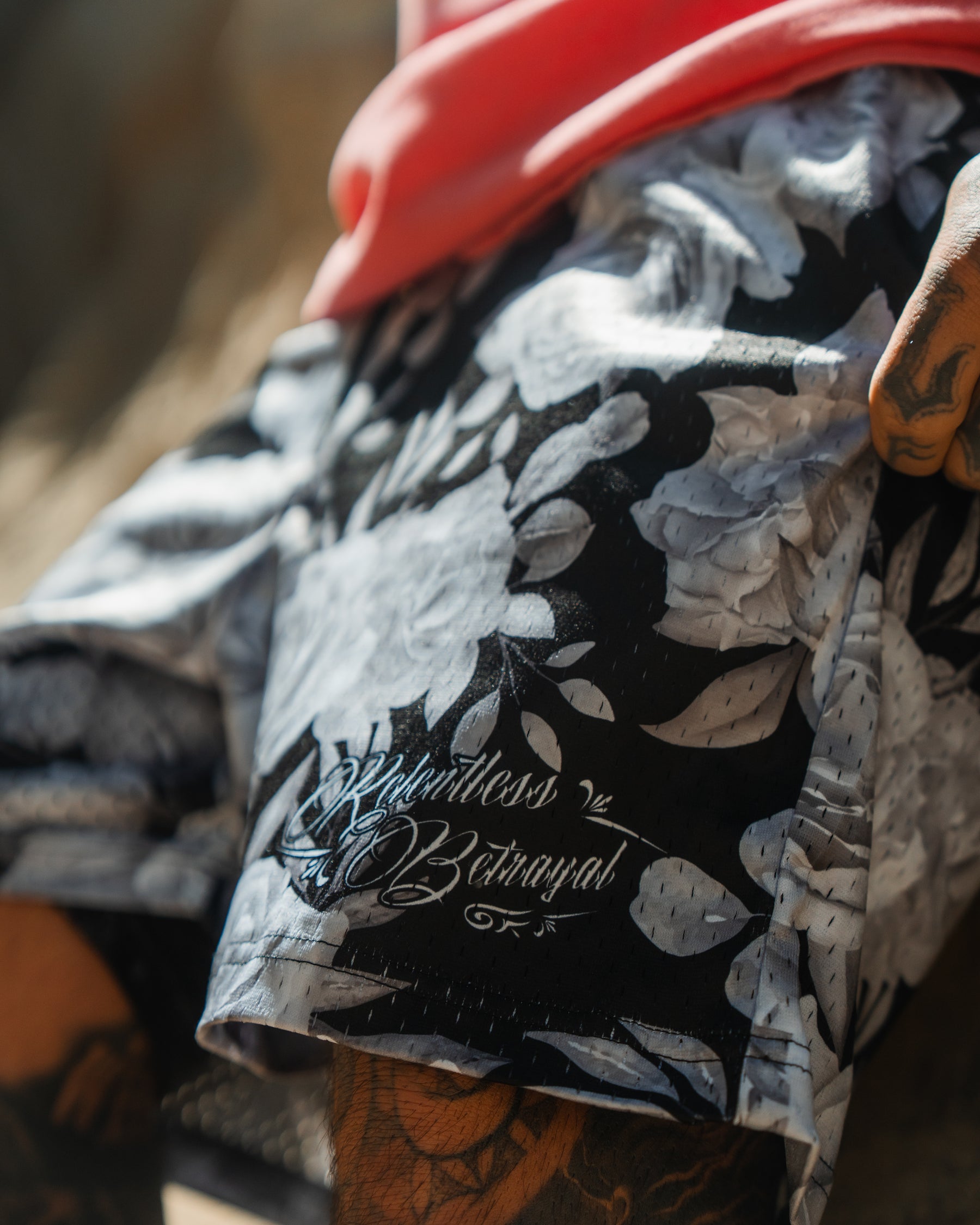 A close-up of a person wearing Relentless Betrayal’s Premium Mesh Shorts | 5- Beauty in Darkness, featuring a black base with white floral pattern and “Boundless Betrayal” script, paired with tattoos for an effortlessly stylish look.